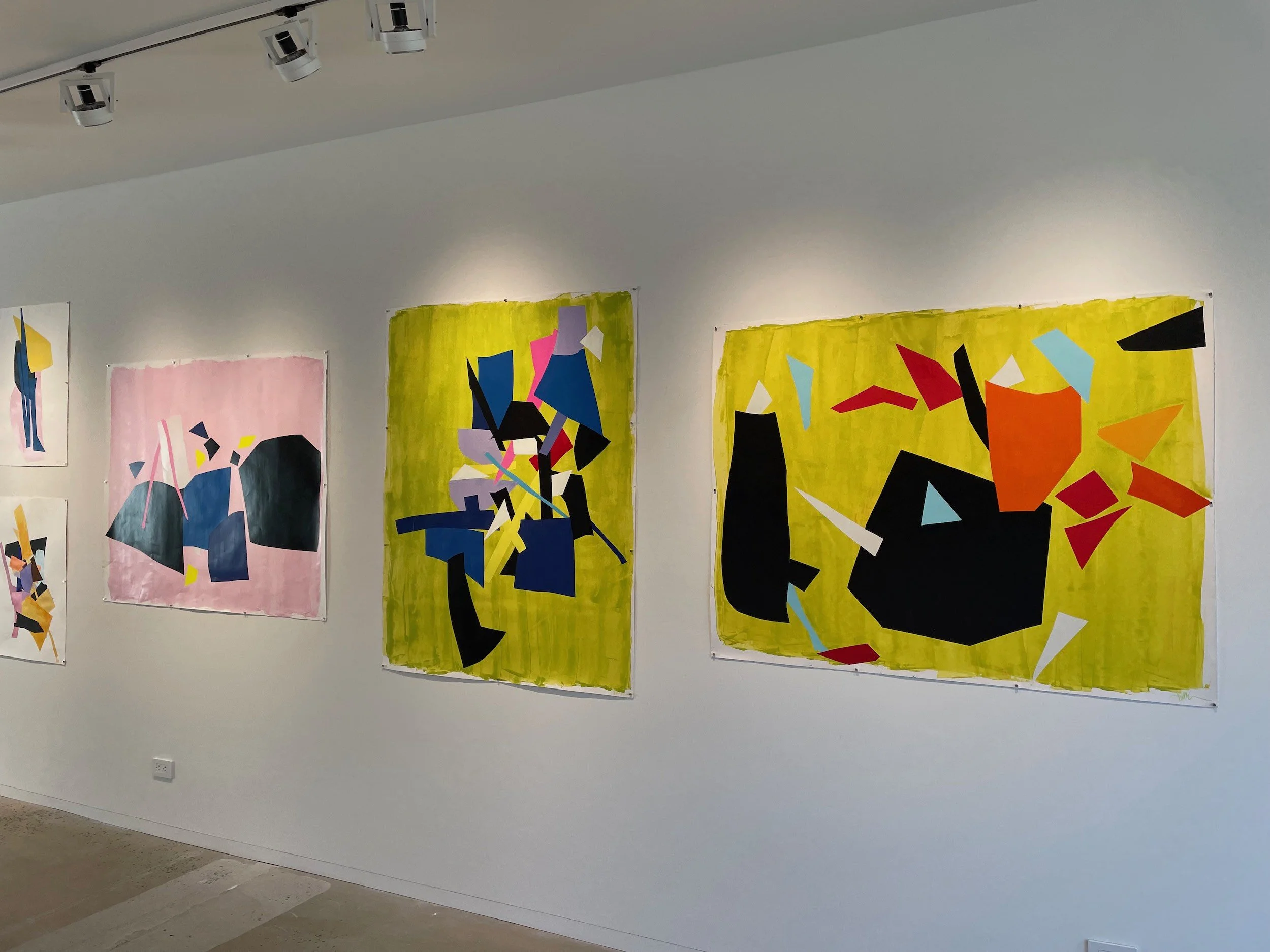 Abstract colorful paintings with geometric shapes hanging on a white gallery wall under directional lighting.