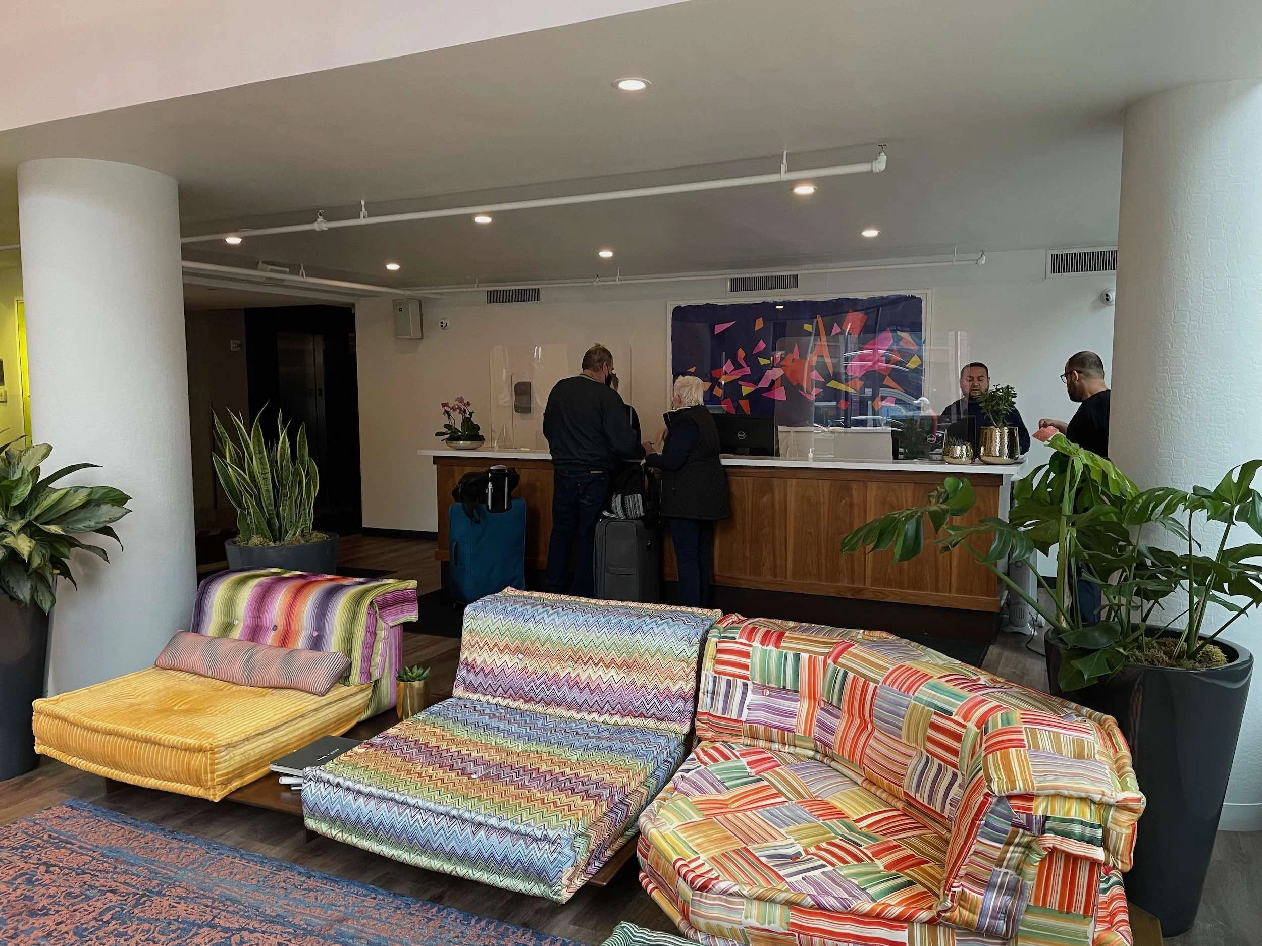 Hotel lobby reception area with colorful patterned couches, green plants in large pots, guests checking in at the counter, and staff behind the reception desk.