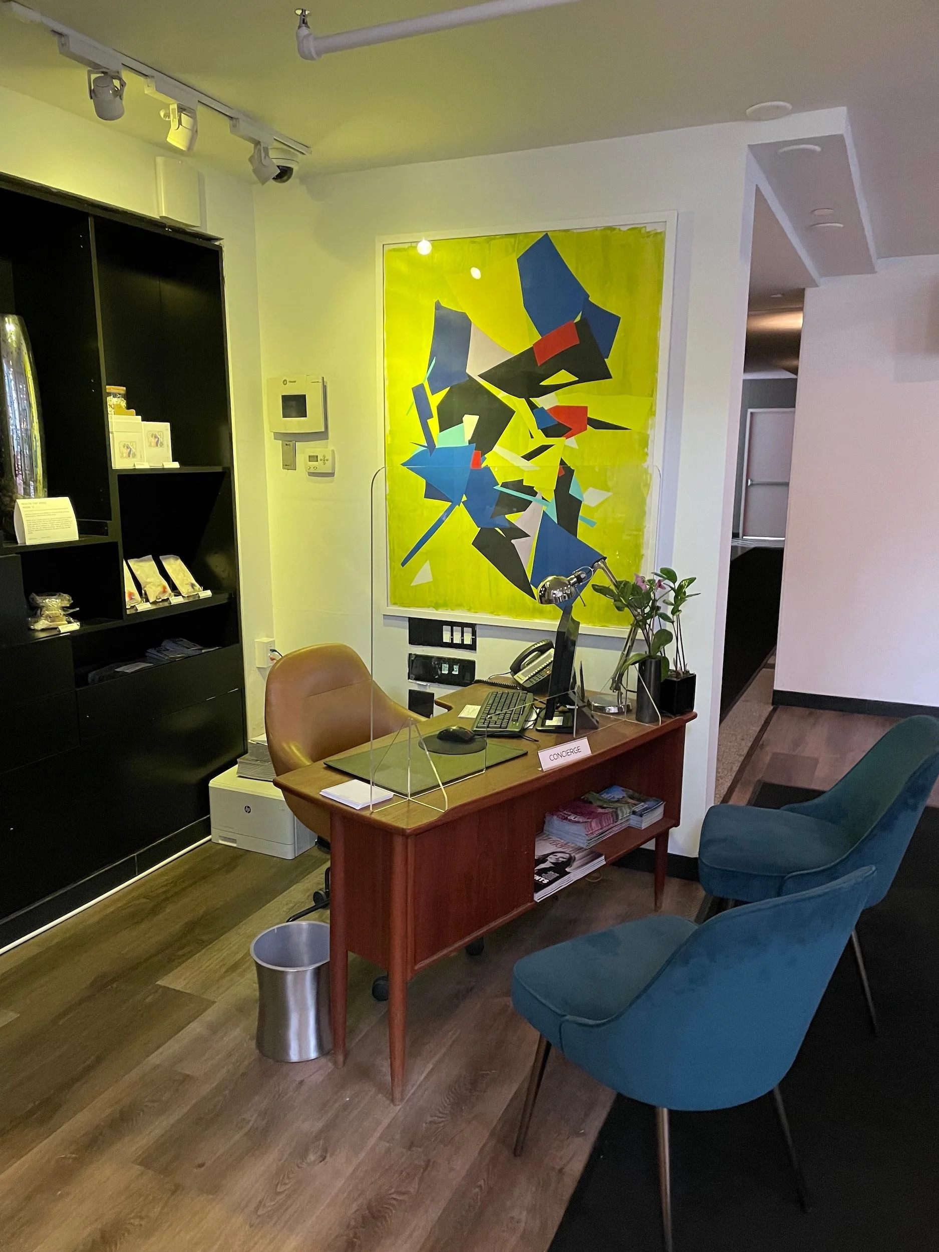 An office reception desk with a computer, a telephone, and potted plants, with colorful abstract artwork on the wall behind it. Two teal chairs are in front of the desk, and a black shelving unit with snacks and a yellow spotlight are visible in the 