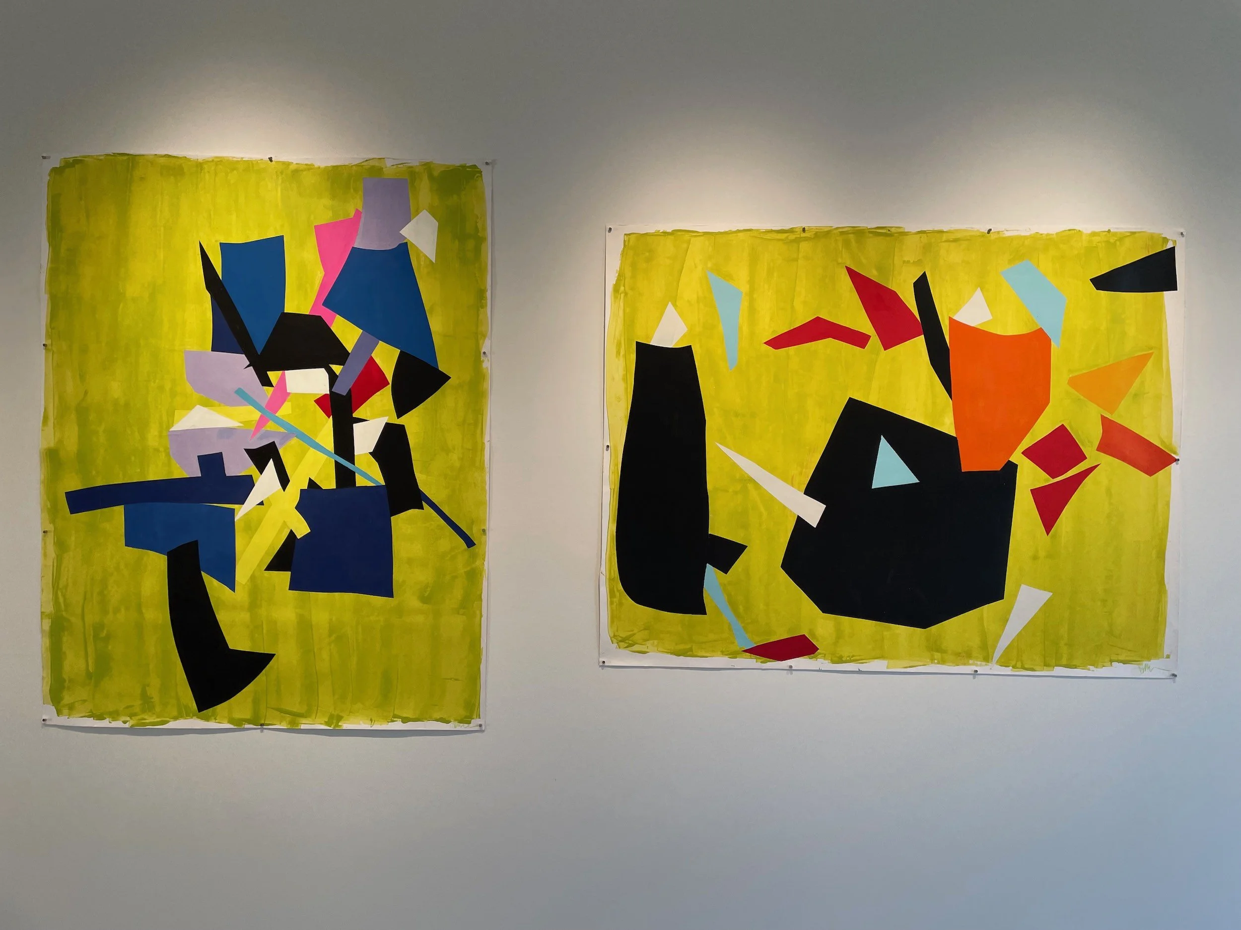 Two abstract paintings with bright yellow backgrounds, featuring various geometric shapes in colors like blue, black, red, pink, and white, hung on a white gallery wall.