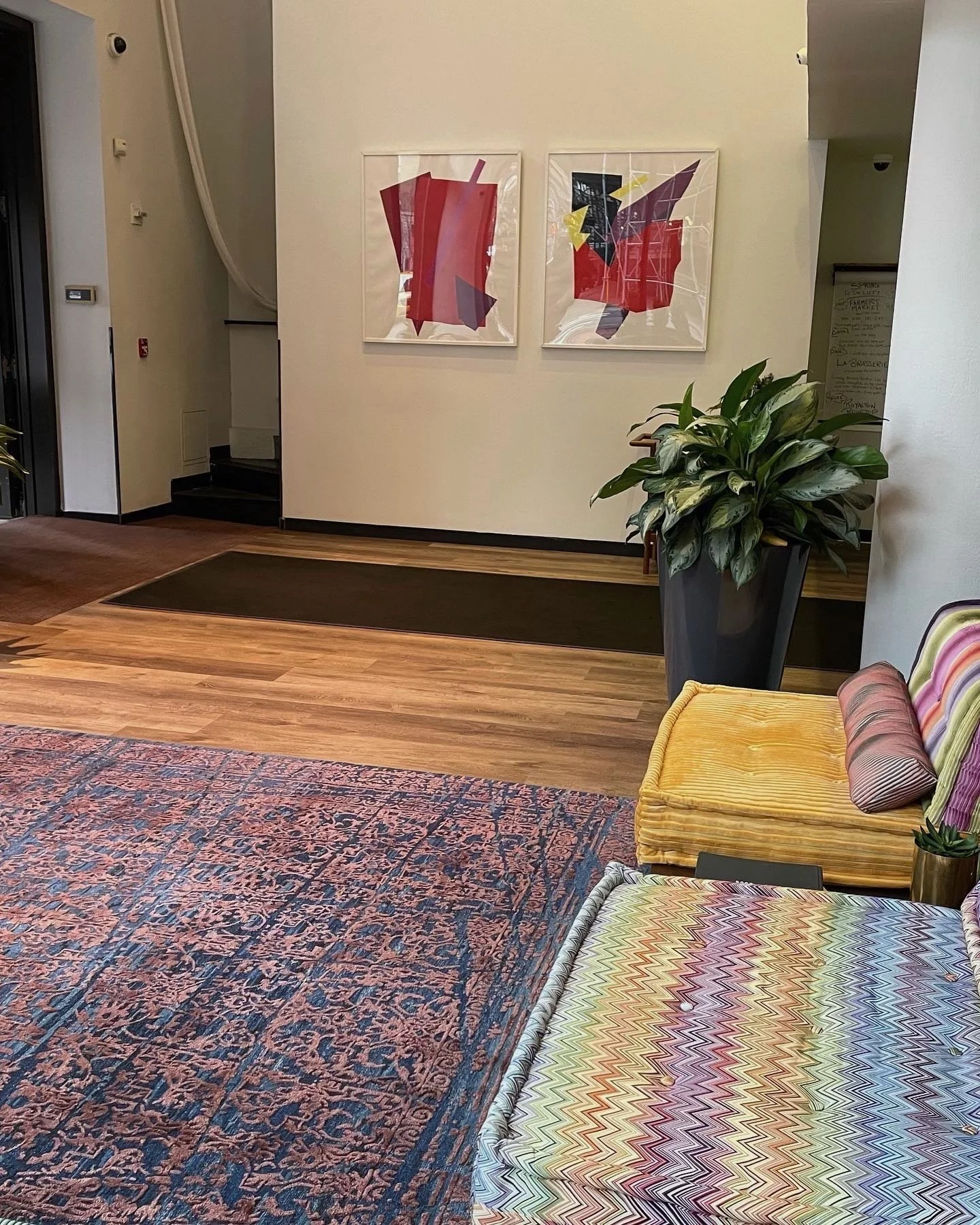 Lobby area featuring colorful abstract artwork on the wall, a large potted plant, a yellow velvet bench, and patterned cushions on a modern chair, with hardwood flooring and a large area rug.