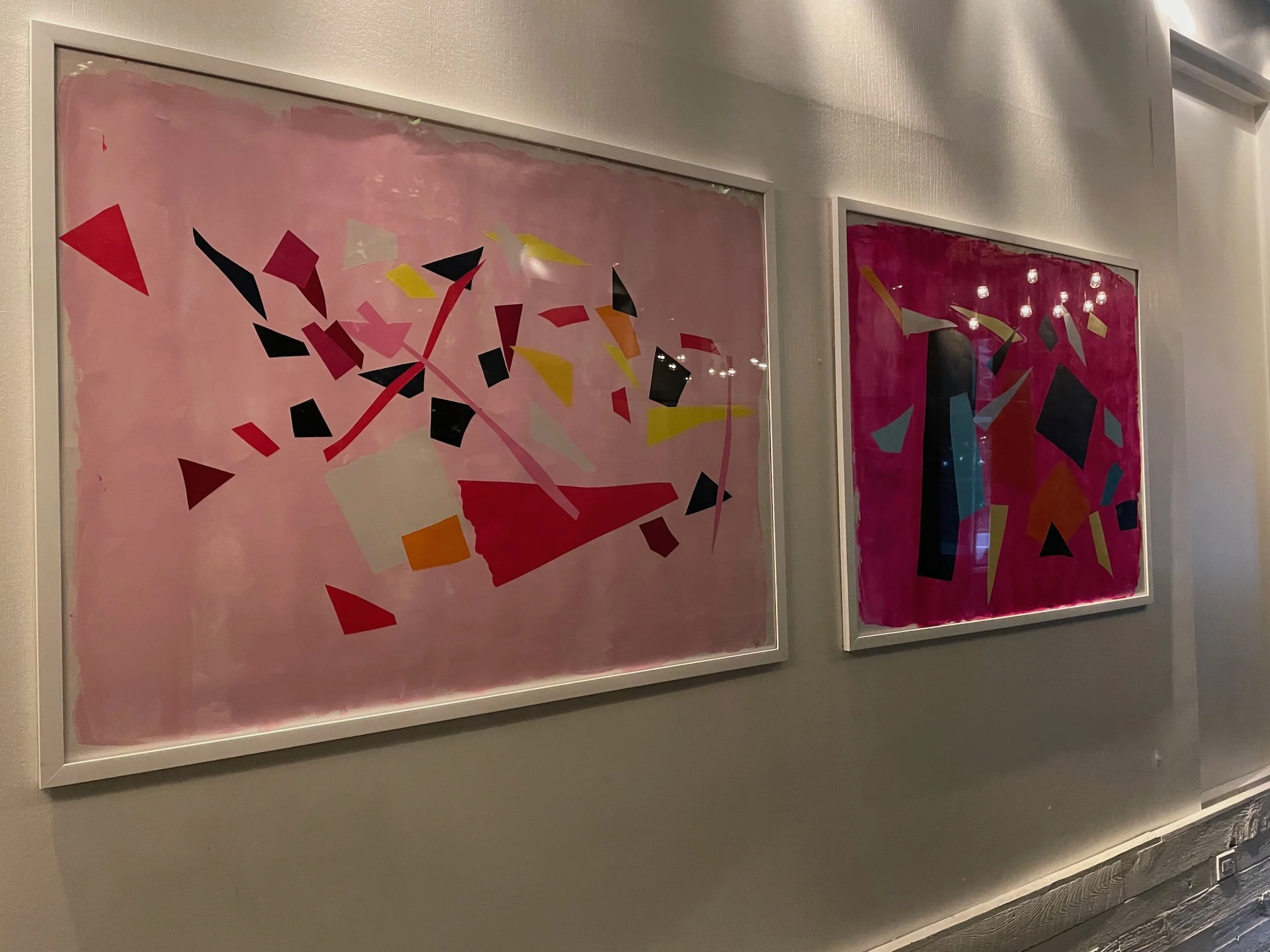 Two colorful abstract art paintings framed and hanging on a light-colored wall, with reflections of ceiling lights visible on the glass.