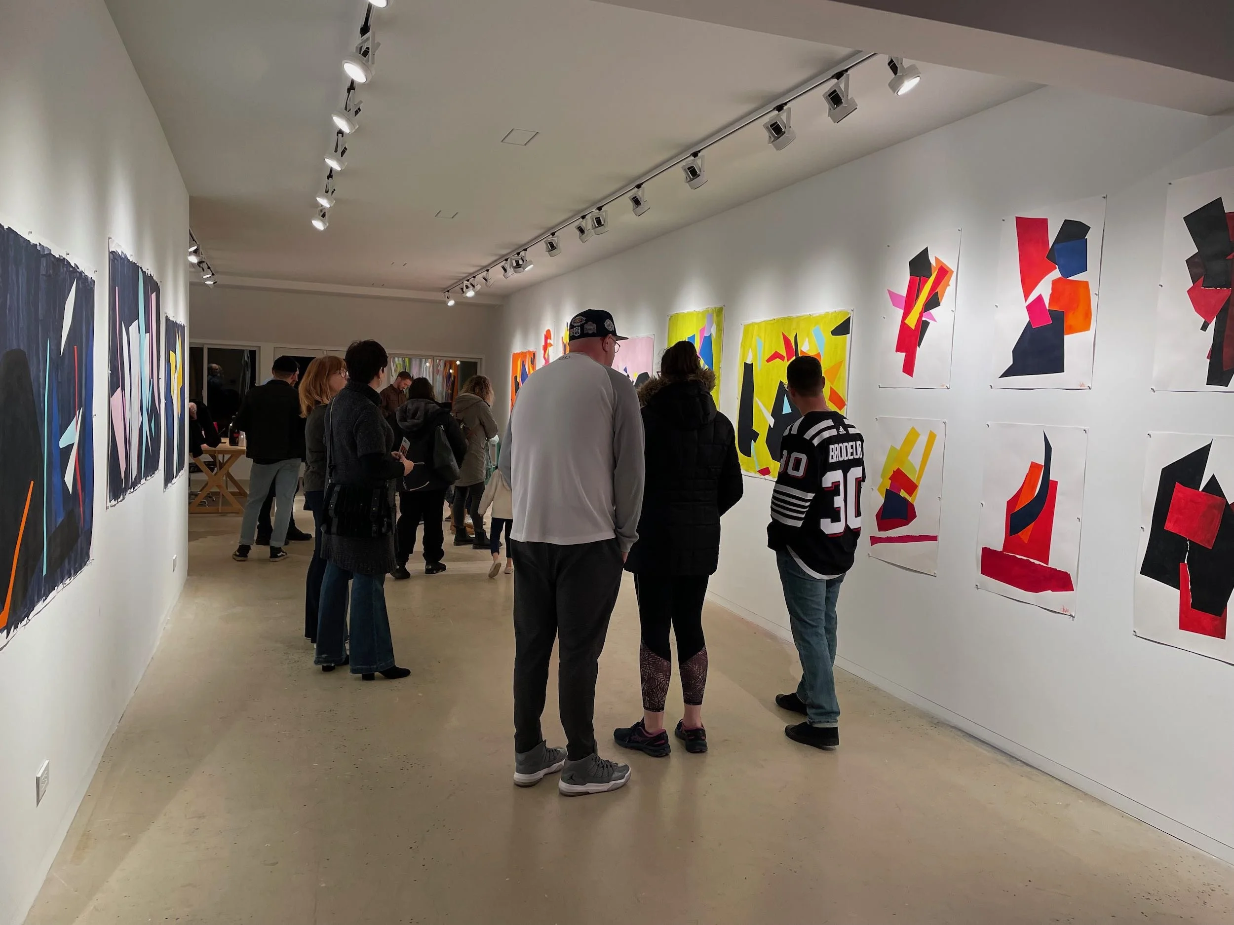 People viewing abstract art paintings displayed on white gallery walls in an art exhibition.