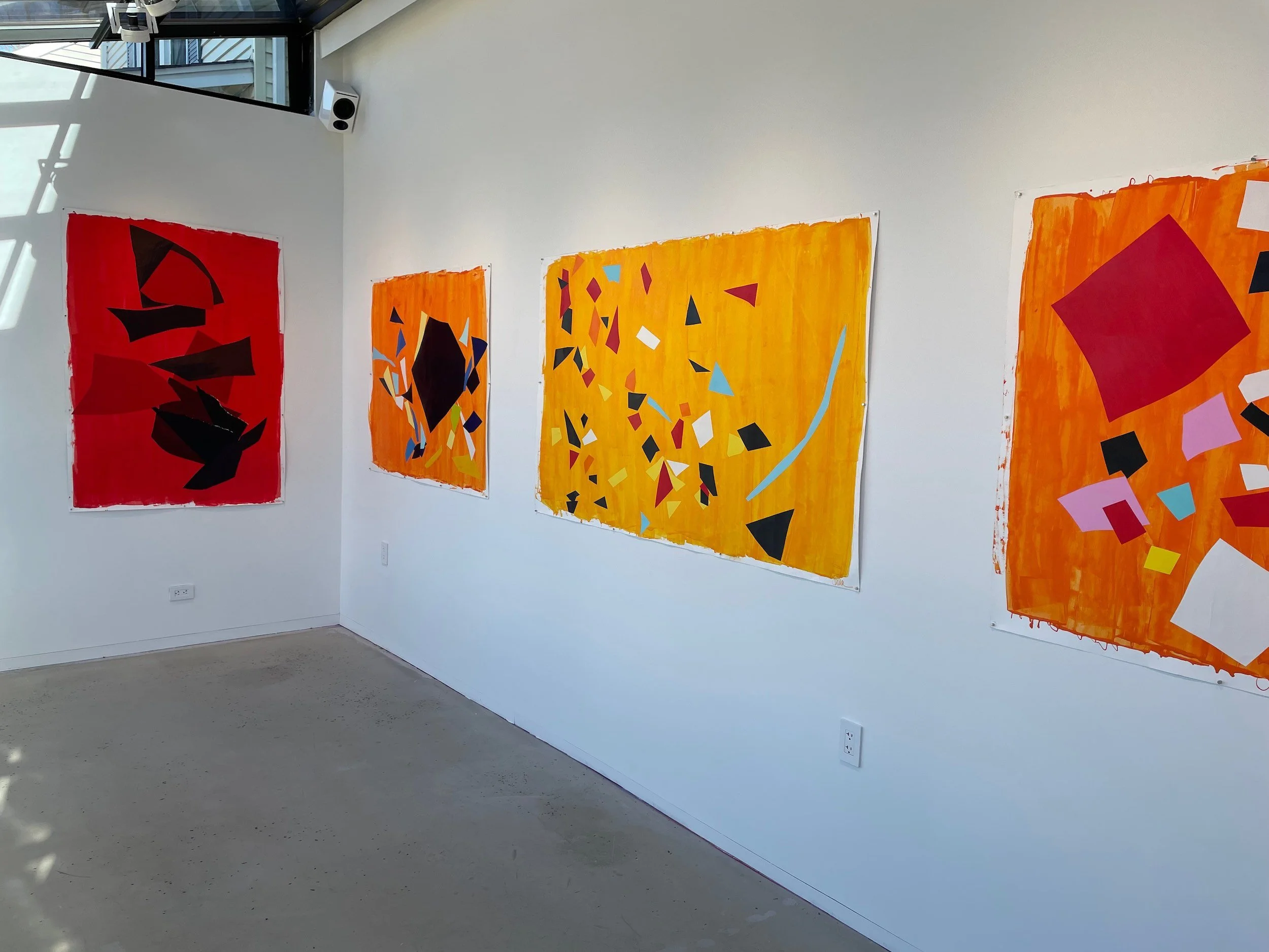 Abstract colorful paintings with geometric shapes on a white gallery wall, featuring reds, oranges, black, pink, blue, and white shapes.