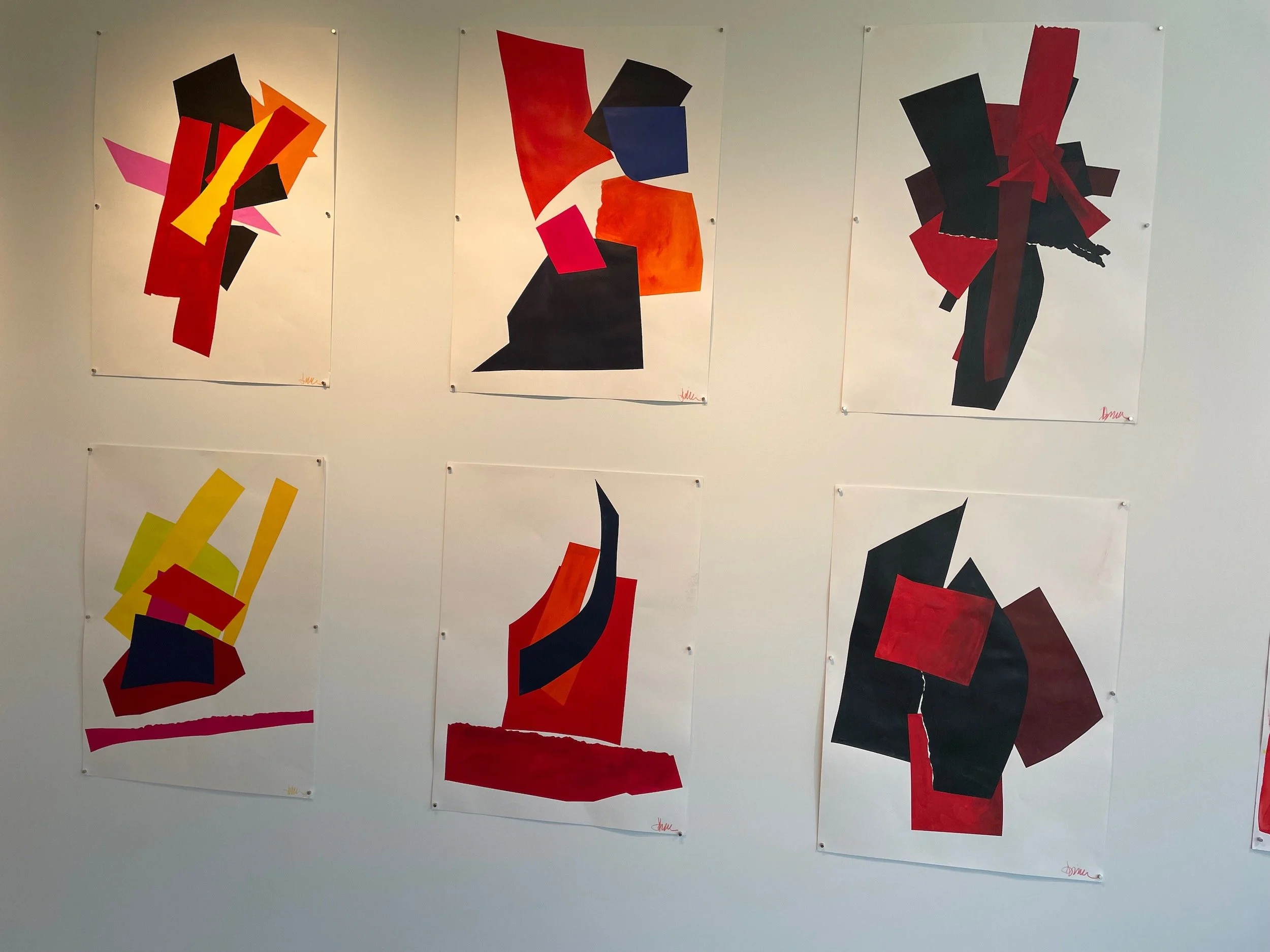 Six abstract collages on white paper, each with bold geometric shapes in red, black, yellow, orange, pink, and blue, displayed on a gallery wall.
