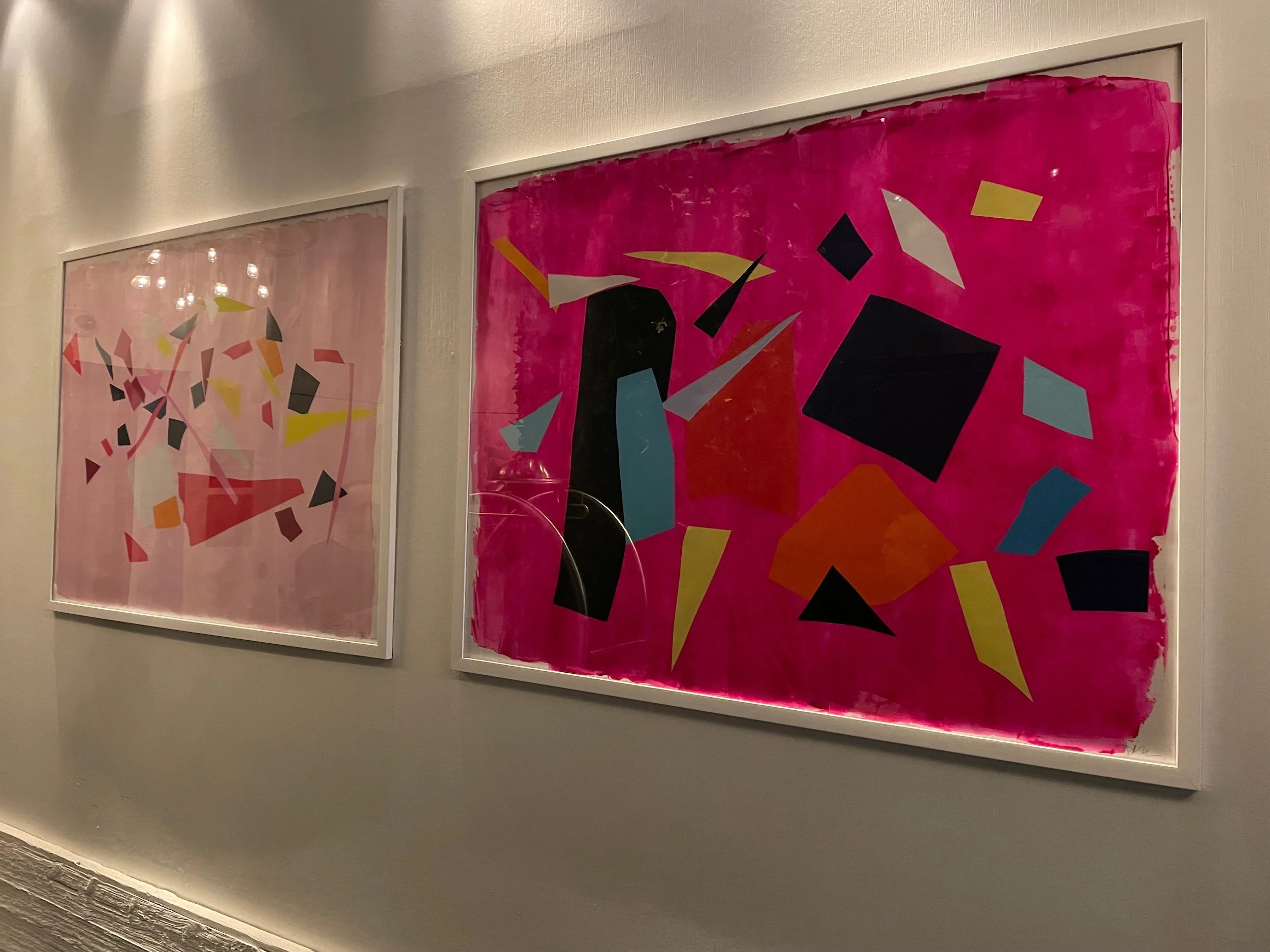 Two abstract geometric paintings with colorful shapes on a pink background, framed and hung on a gallery wall.