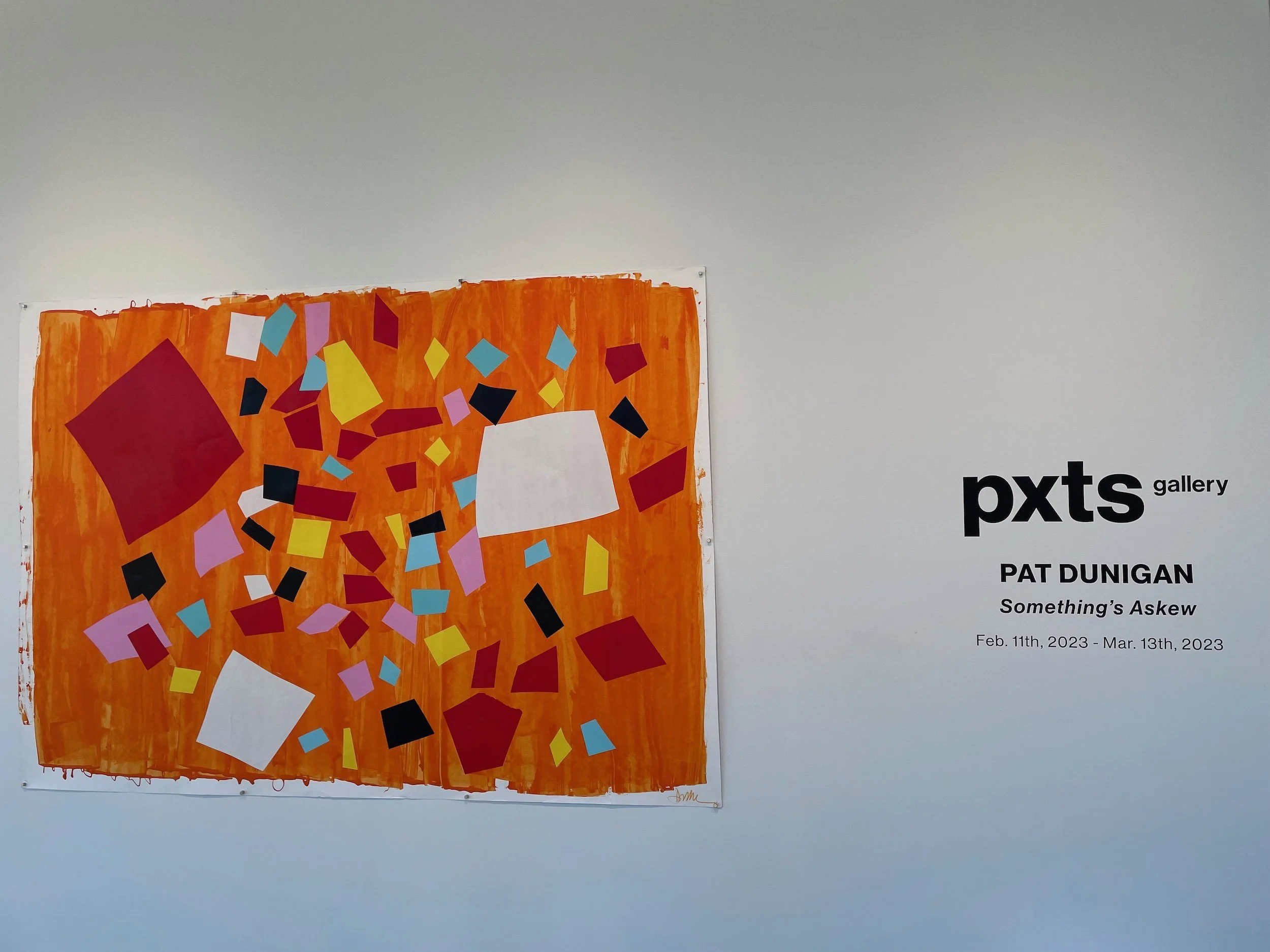An abstract artwork with multicolored geometric shapes on an orange background, displayed in an art gallery. Next to the artwork is a sign that reads 'pxts gallery PAT DUNIGAN Something's Askew Feb. 11th, 2023 - Mar. 13th, 2023.'