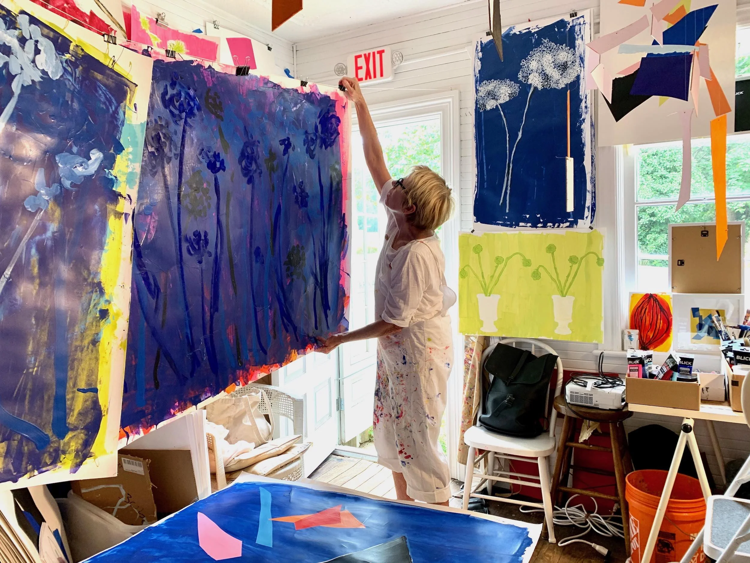 An artist with short blonde hair and glasses is working on a large abstract painting in a bright studio. She is wearing a white shirt and paint-splattered pants, reaching up to paint or hang something on the wall. The studio is filled with colorful abstract artworks and creative supplies, with windows letting in natural light.