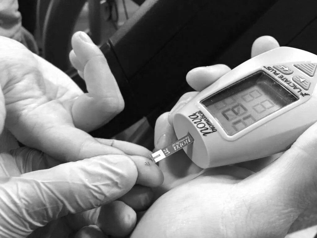 A healthcare worker, wearing gloves, is using a lancet to prick a person's finger for blood glucose testing. The person’s hand is extended, and the lancet device is positioned to draw blood. A glucose meter and test strip are also visible.