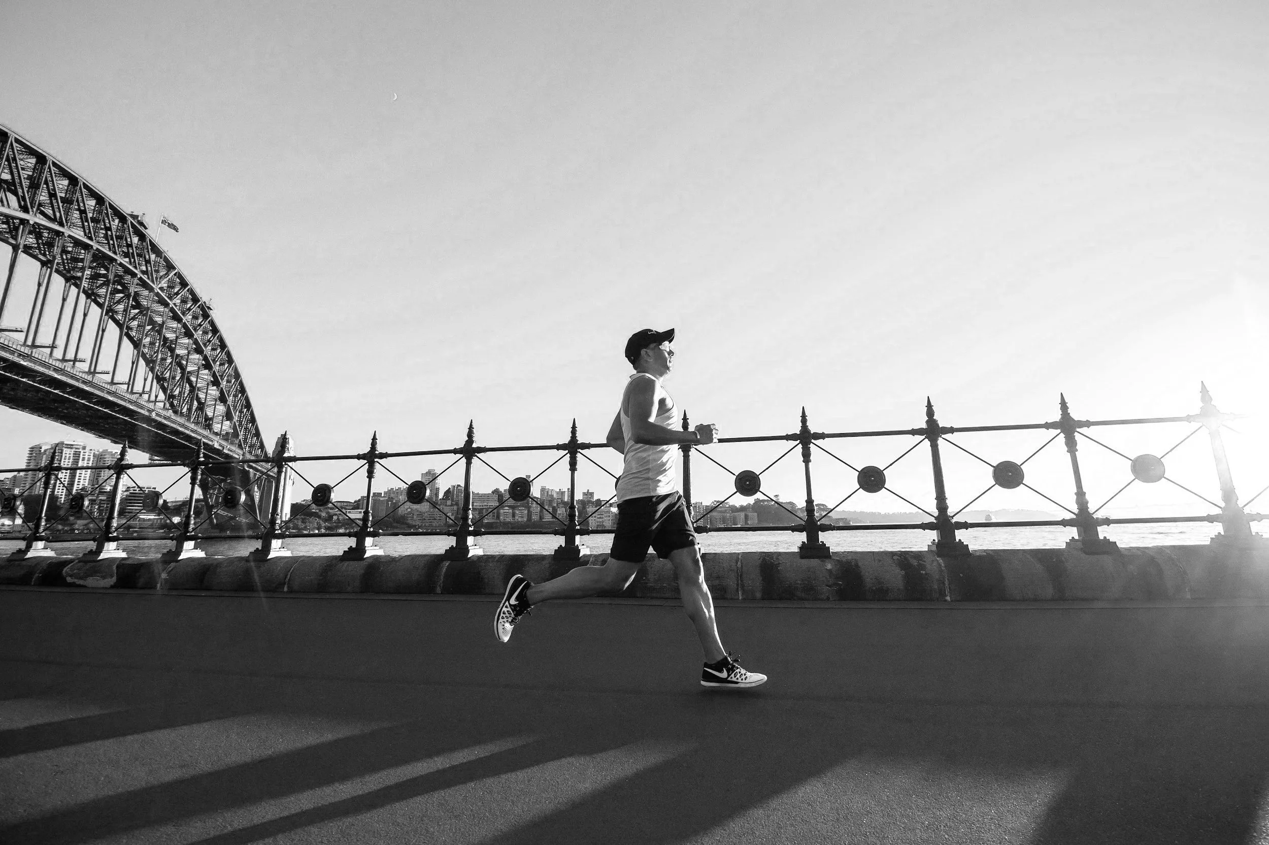 A person jogging along a waterfront promenade with a bridge and cityscape in the background, black and white photo.