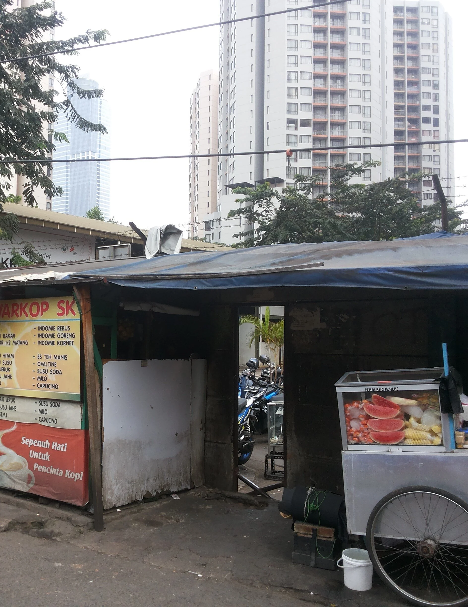 Figure 4. A wall divides Kampung Kuningan from its upper-middle class neighbor, while a cut reveals how interconnected the two settlements in fact are, 2015. The tower’s developer built the wall in order to increase the value of the apartments. Yet, many residents of Kampung Kuningan work in the high-rise, as drivers, maids, gardeners, handymen, security personnel, and vendors, while other employees, who live further afield, may take advantage of Kuningan’s food stalls from work. Jakarta is filled with similar wall and cuts. Photograph by Abidin Kusno.