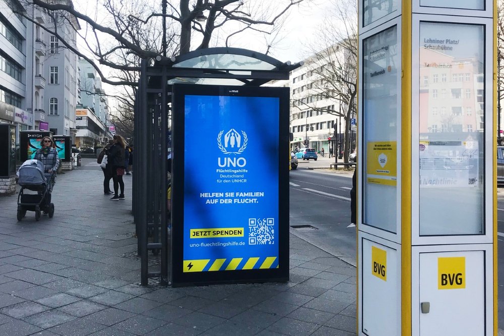  Abbildung 12. Digitale Werbung an einem Buswartehäuschen, Berlin, Deutschland, März 2022. Foto von Mireille van der Moga.  Die digitale Anzeige macht das Spenden für das UNHCR, das Flüchtlingshilfswerk der Vereinten Nationen, einfach, indem sie eine