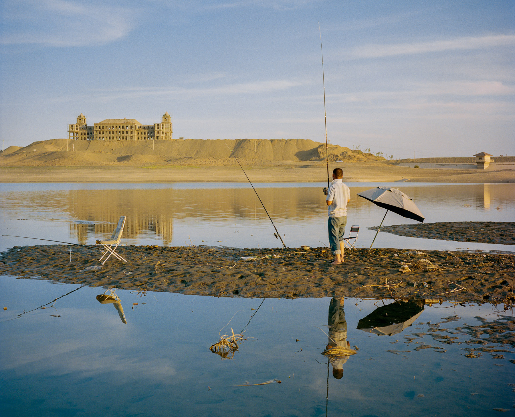  June 2016. Xinjiang province, China. A Han Chinese migrant worker fishing in a small lake in the outskirts of Turpan. In the background, a typical item of China's fast developping West, a half-built half-decaying palace or government building. 