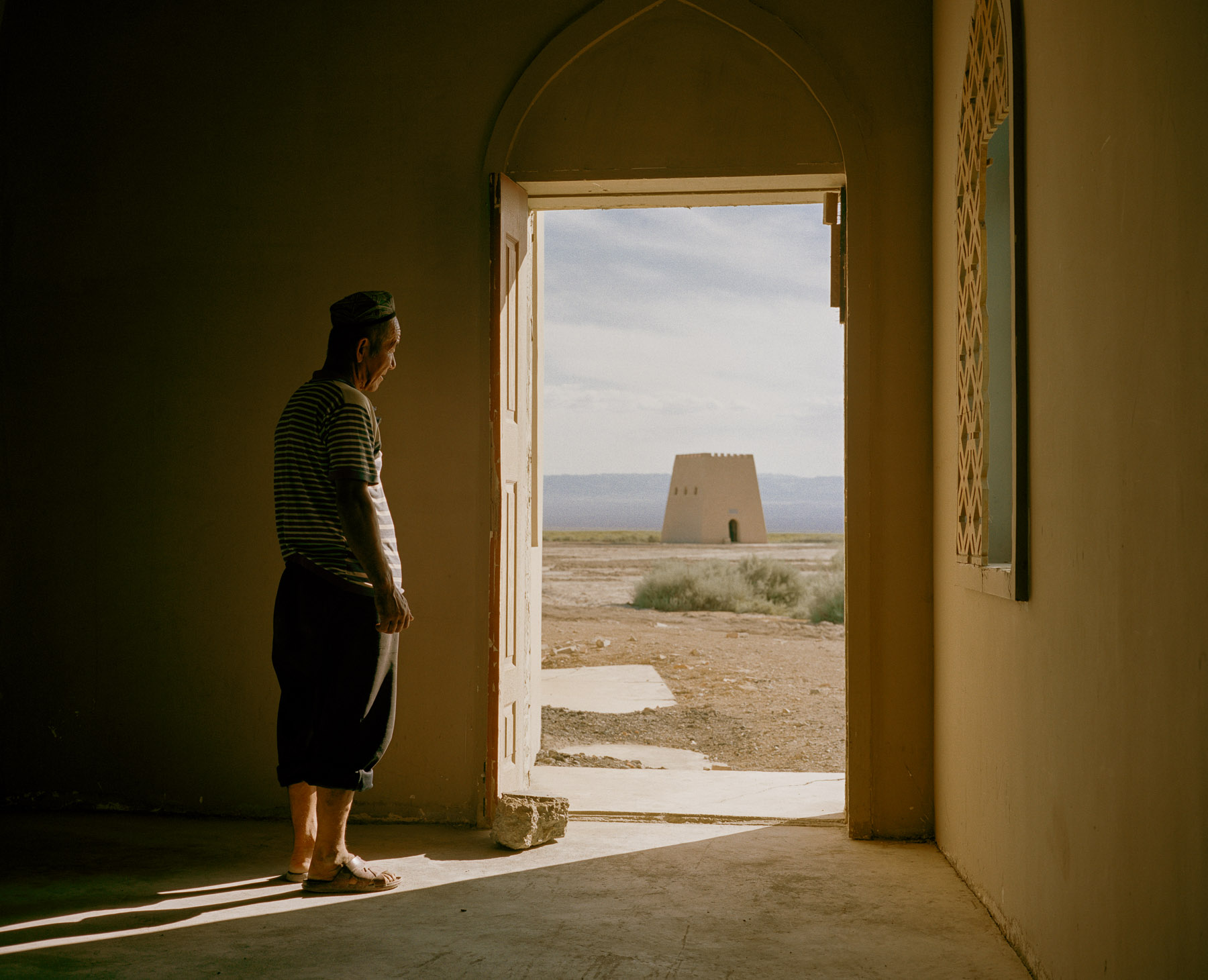 June 2016. Xinjiang province, China. Uighur man looking through the door of his house into the Turpan depression and Ayding lake. Ayding lake is a dried up lake in the Turpan Depression, Xinjiang Uyghur Autonomous Region.. At 154 m below sea level, 