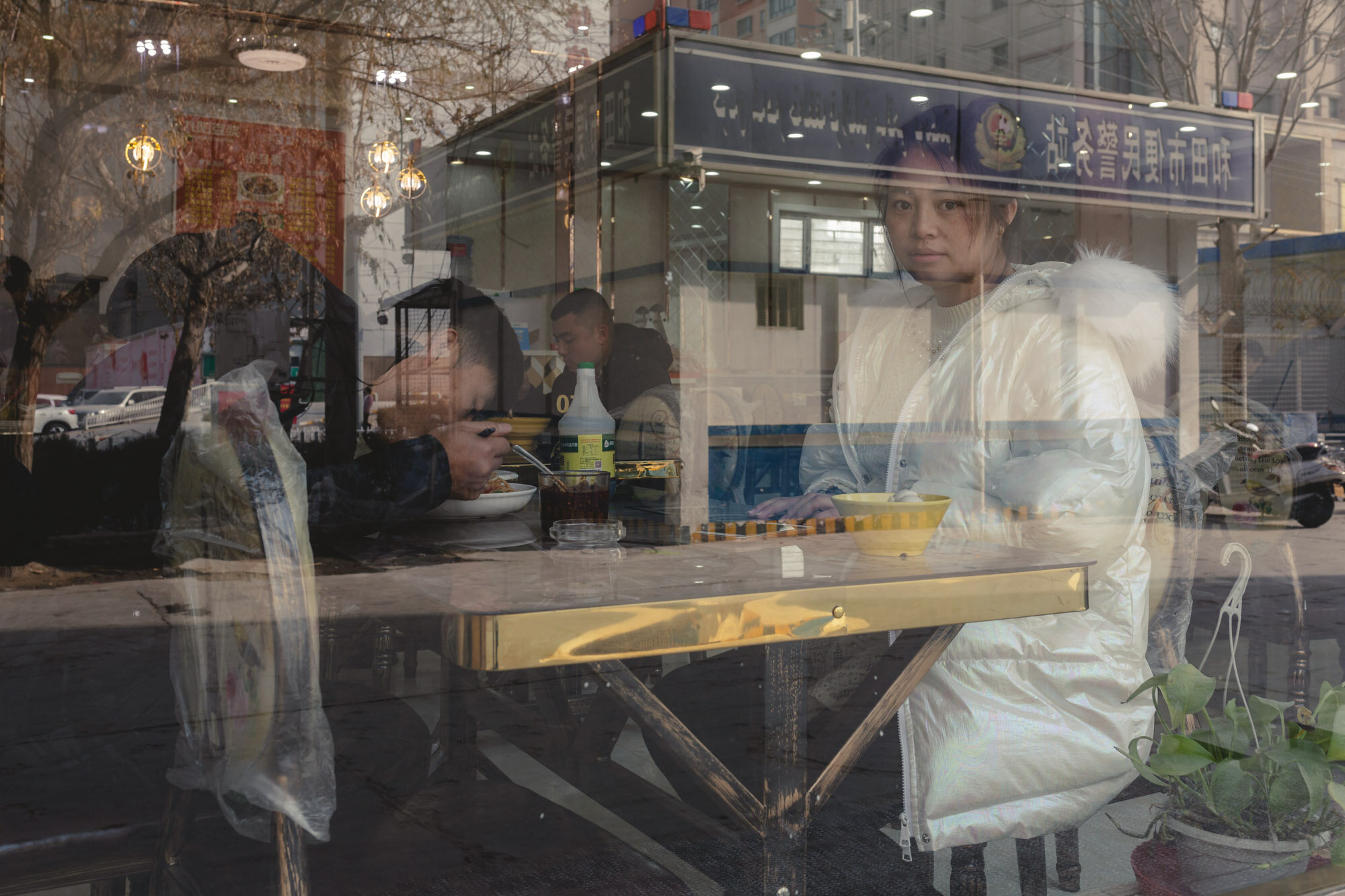  February 5th 2019. Hotan, Xinjiang province. Couple eating lunch in a local restaurant. In the reflection in the window one of the many additional police stations that have been installed at almost every street corner over the past three years. 