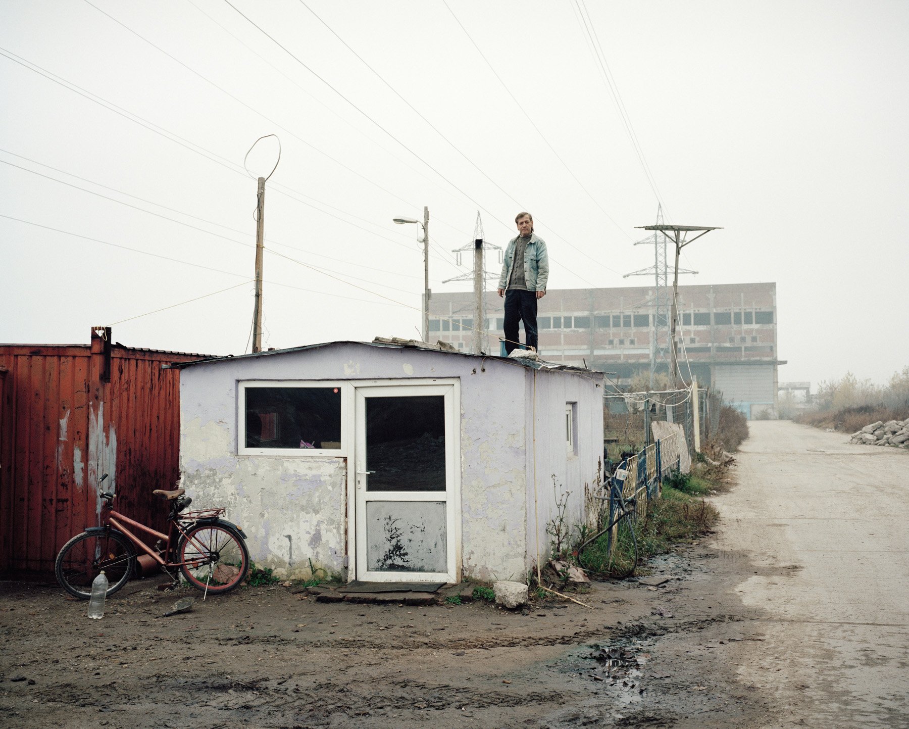  Romania, Giurgiu. 2016. A man working as security guard stand on a barrack in front of an abandoned factory that he watch. 