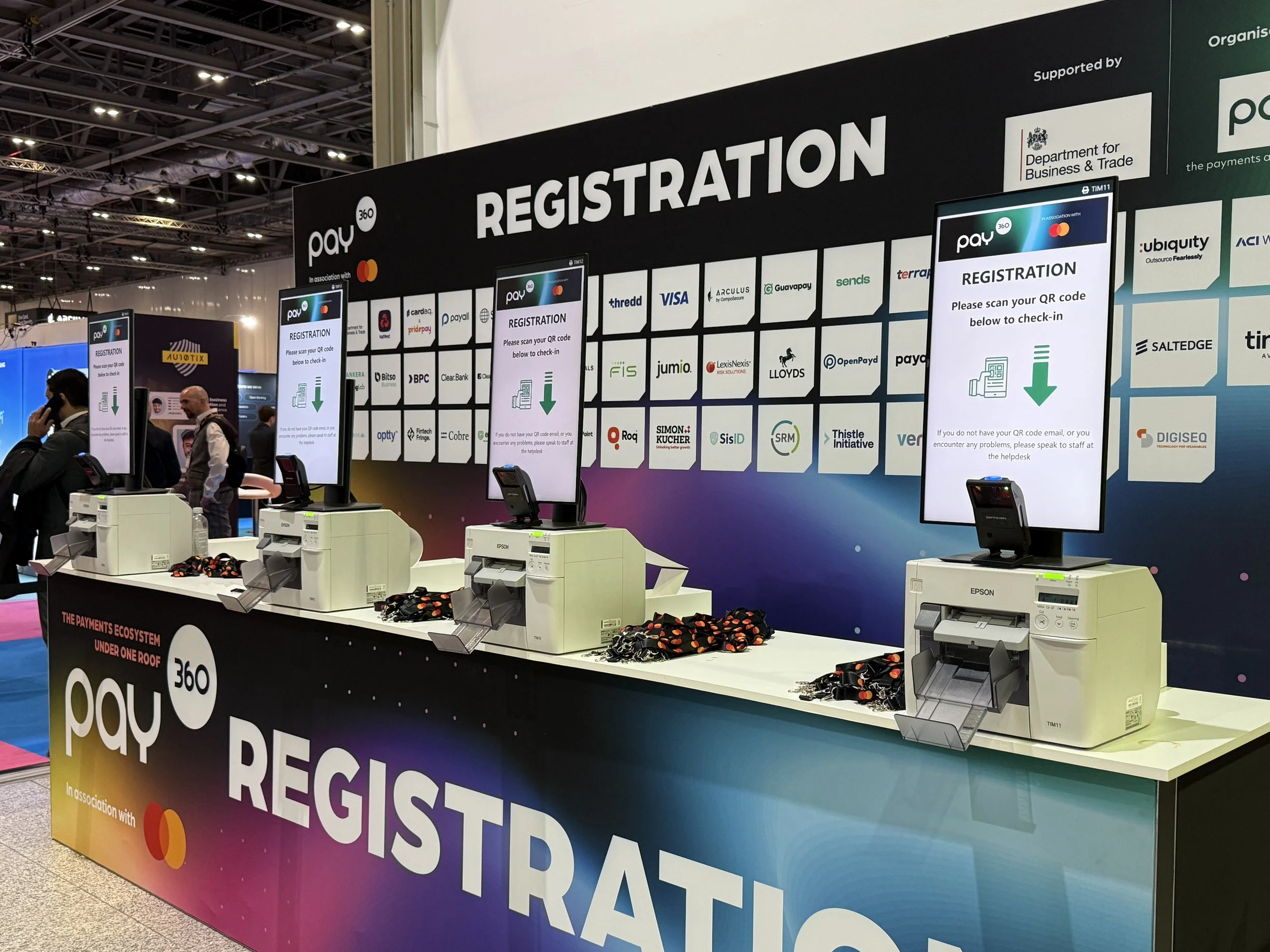 Self scan badge printing machines also know as kiosks are lined up along a branded registration desk. Lanyards are placed next to each badge printer and signs are hanging behind each badge printer /kiosk telling event visitors to scan their QR code.