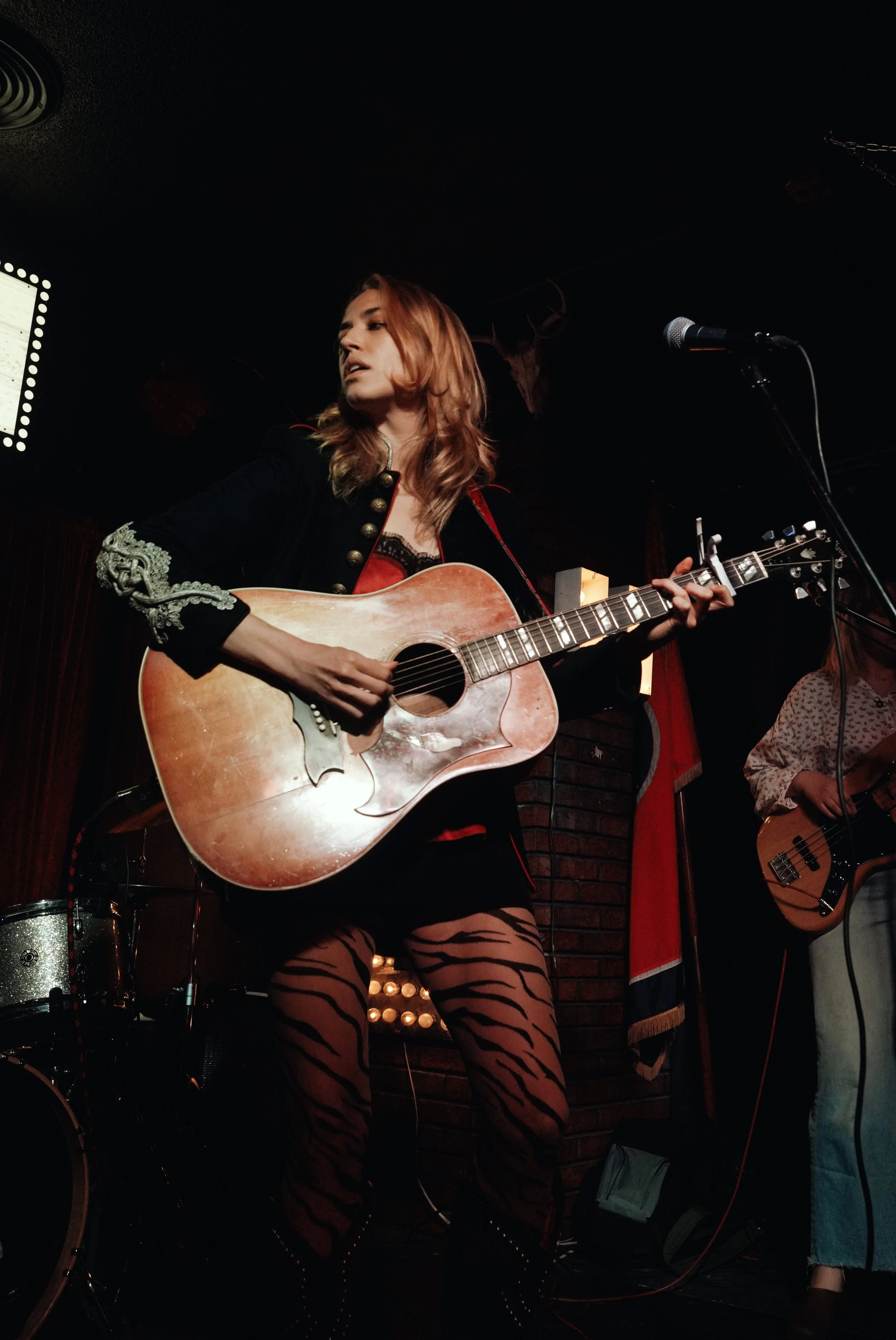 A woman with red hair is playing an acoustic guitar on stage, wearing a black outfit with ornate silver embellishments and tiger-striped stockings, in a dimly lit venue with a microphone nearby.