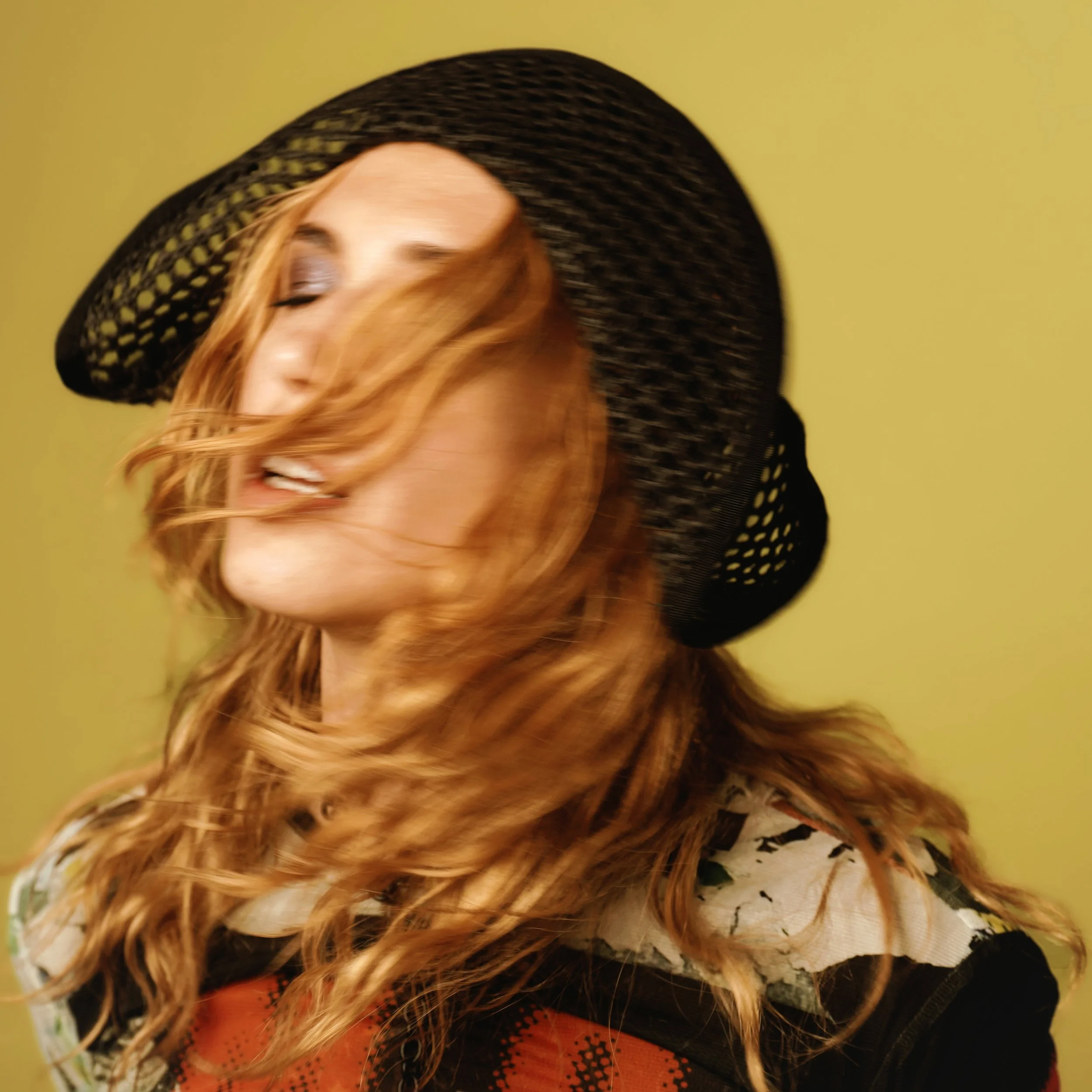 Americana artist Melanie MacLaren with long, curly red hair wearing a black, wide-brimmed hat with an open weave pattern, looking down against a yellow background.