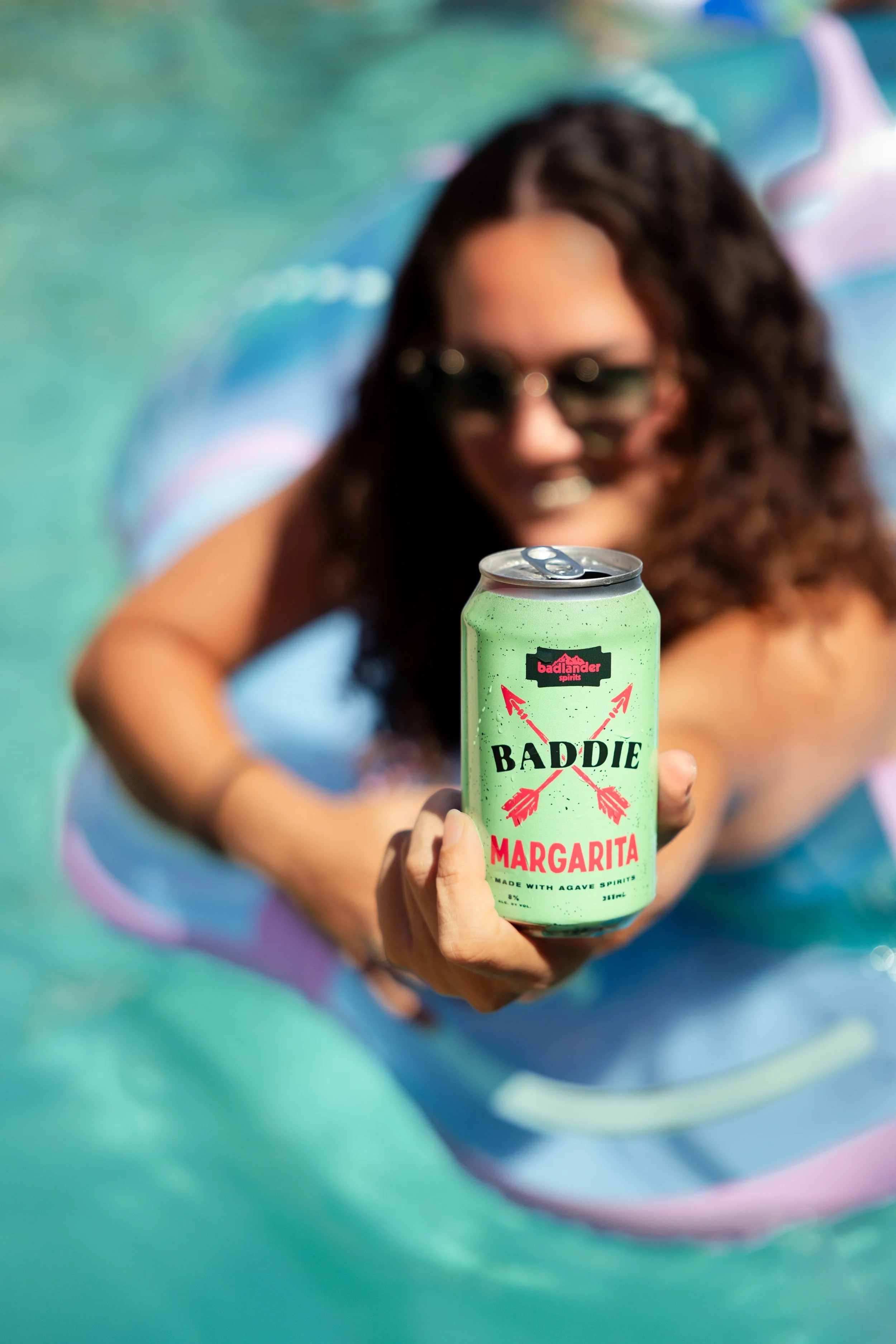 A woman with curly hair and sunglasses is holding a green can of Badlander Spirits Baddie Margarita in front of her, with her other arm resting on a colorful, blurred background.