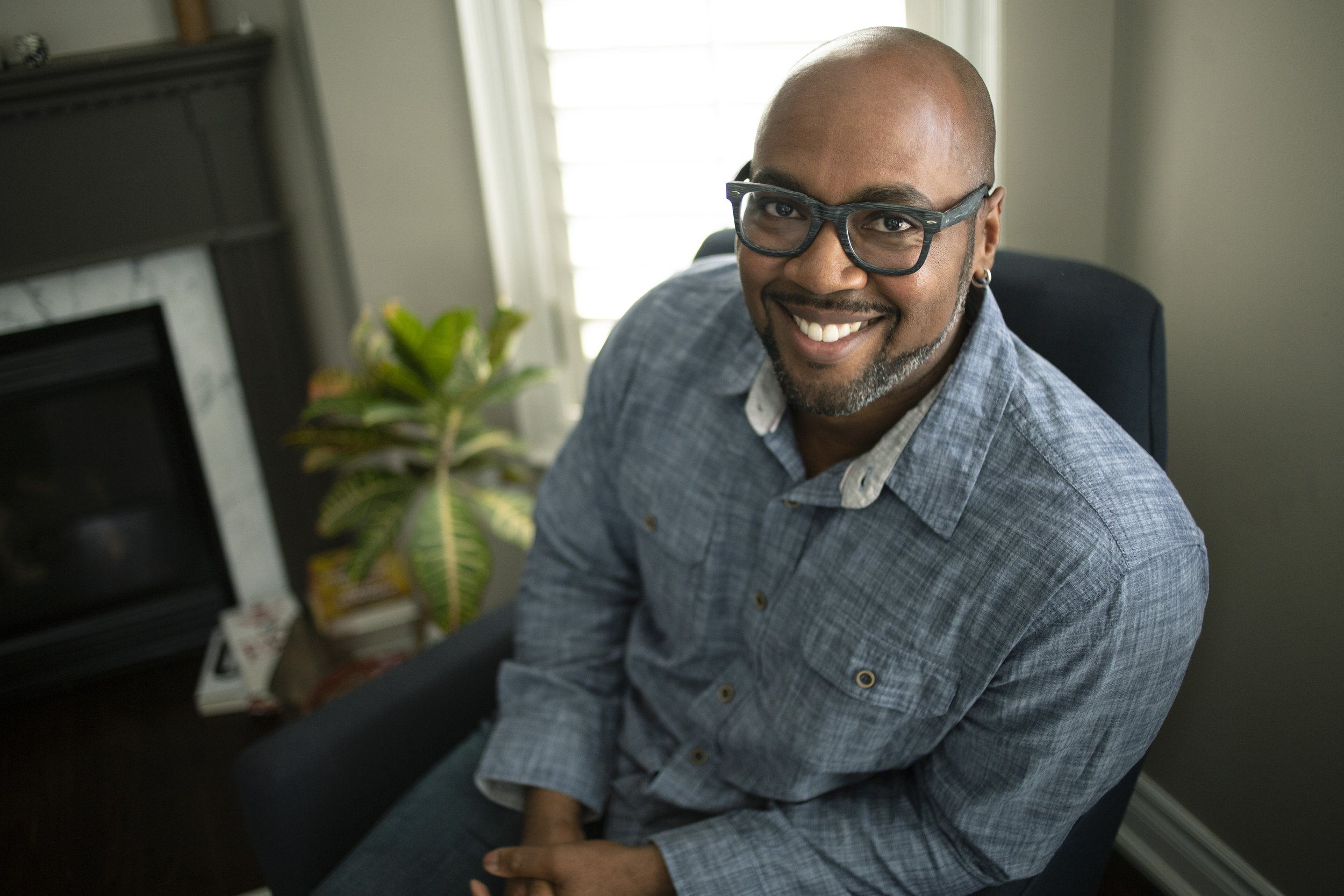 A smiling bald man with glasses wearing a gray checkered shirt, sitting in a room with a window, a plant, and a fireplace in the background.