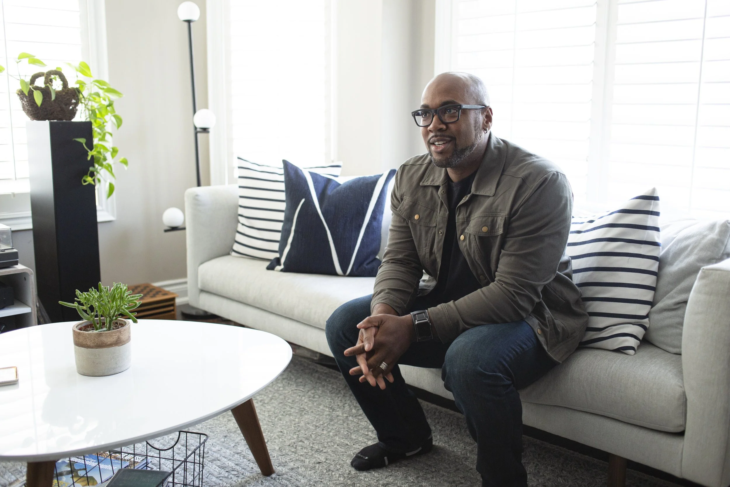 A man with glasses and a beard sitting on a sofa in a bright living room, with a plant on a side table and striped pillows behind him.