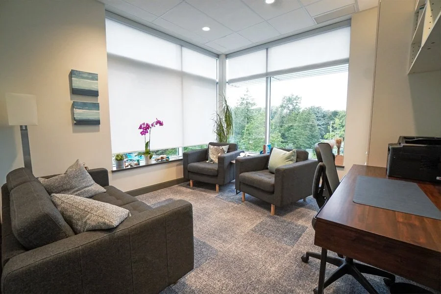 Modern office lounge with gray sofa, two gray armchairs, and large windows with view of trees outside