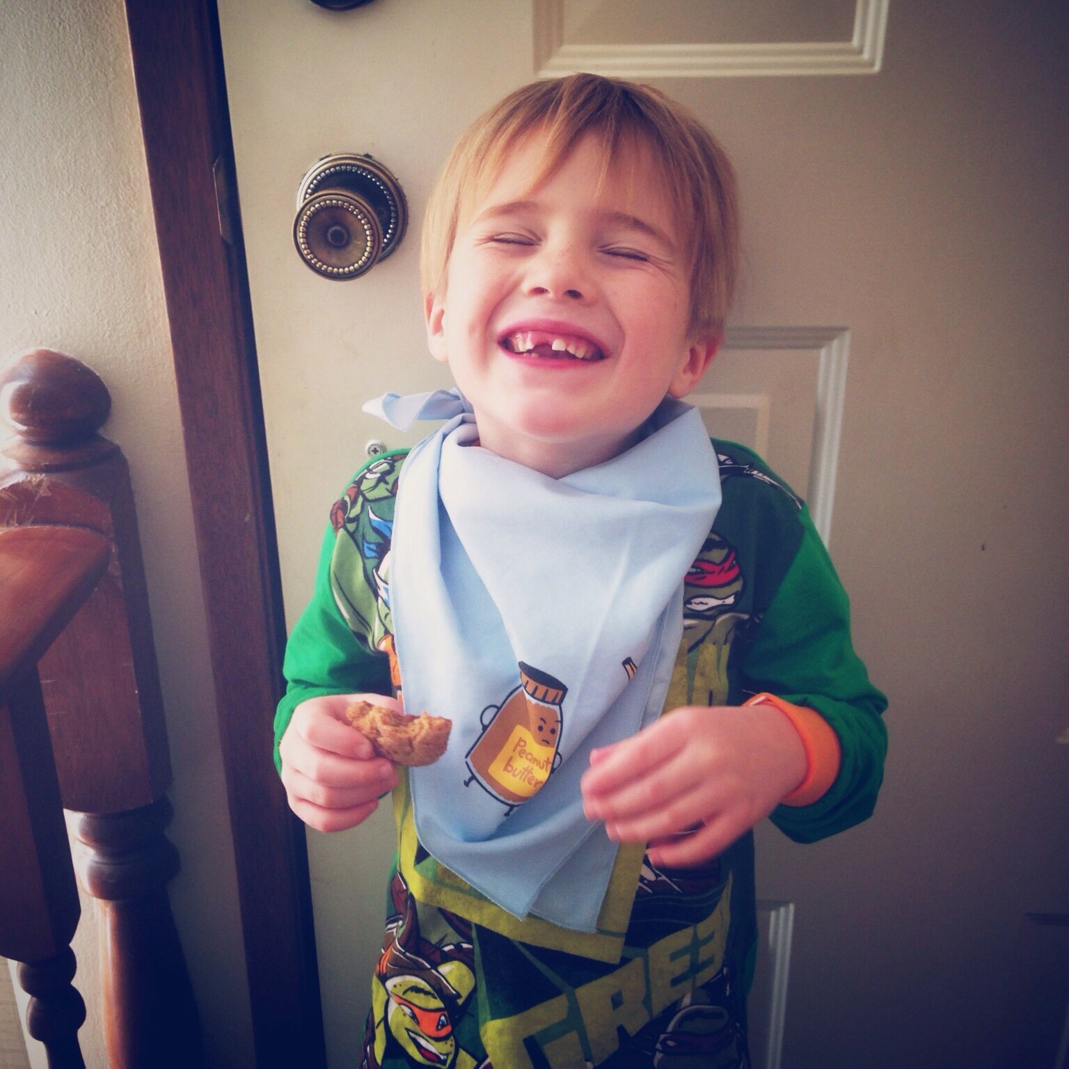   PB vs. Banana napkin/scarf.  Photo of a little boy without a front tooth laughing as he eats a cookie, with a light blue napkin tied around his neck. The napkin illustration is of a jar of peanut butter and banana facing each other, with hands on t