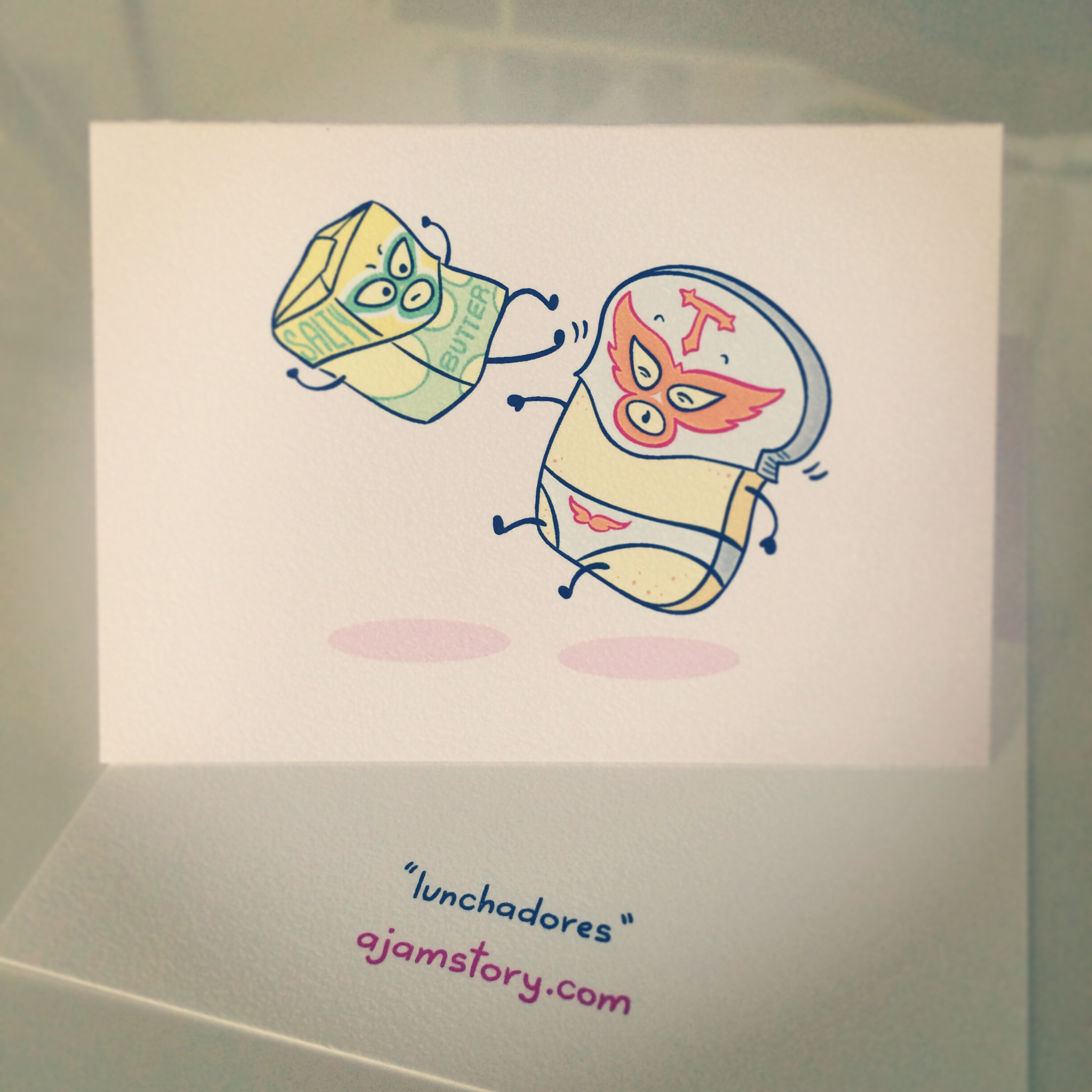   “Lunchadors” : A photo of a small greeting card depicting an illustration of Butter and Toast dressed as Mexican luchadors, with butter in the air kicking, and toast falling backwards. The back of the card says "lunchadors” and “ajamstory.com”. 