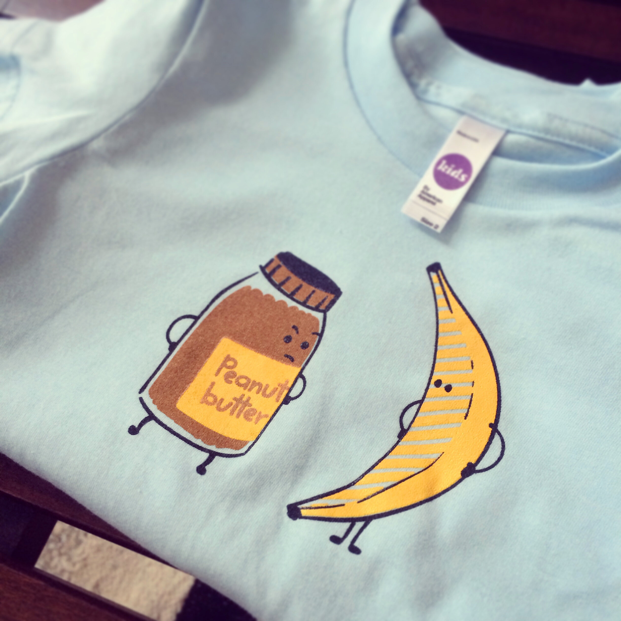   PB vs. Banana tshirt.  A folded light blue children’s t-shirt with an Illustration of a jar of peanut butter and banana facing each other, with hands on their hips. 