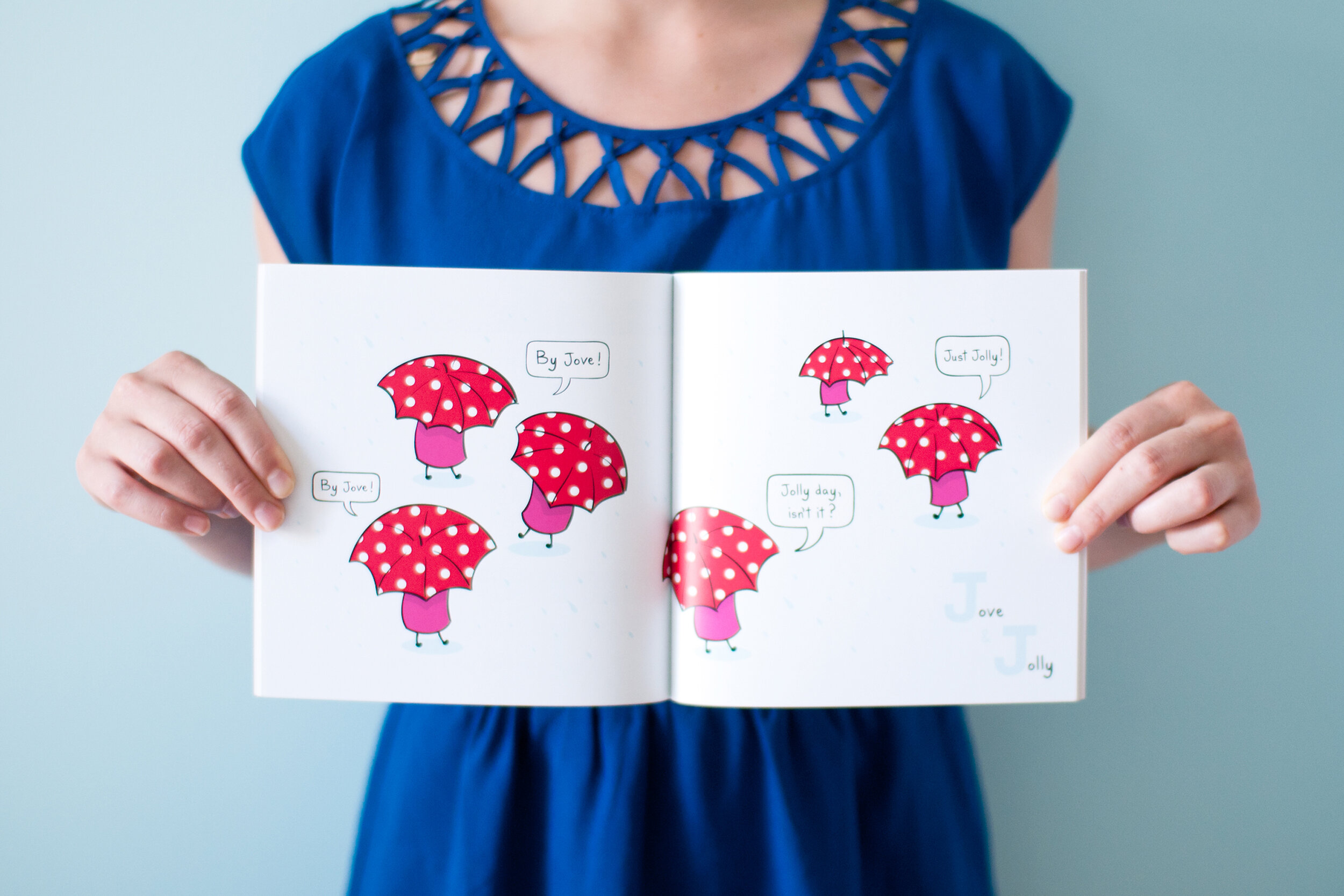  Cropped body of woman wearing a blue dress holding open  A Jam Story: J is for JAM  children’s book in front of her. The book pages display a scene where 6 jars of jam are wandering around with red polka dot umbrellas, saying “By jove!” and “Jolly d