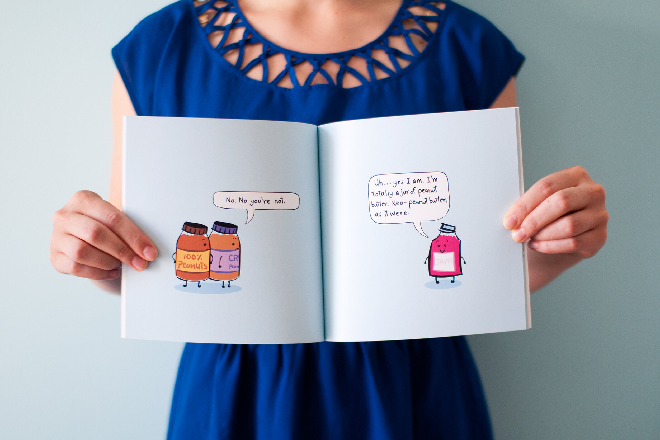  Cropped body of woman wearing a blue dress holding open  A Jam Story  children’s book in front of her. The book pages display a scene where two jars of peanut butter are saying “No, No you’re not.” to a jar of jam, who is replying “Uh… yes I am. I’m