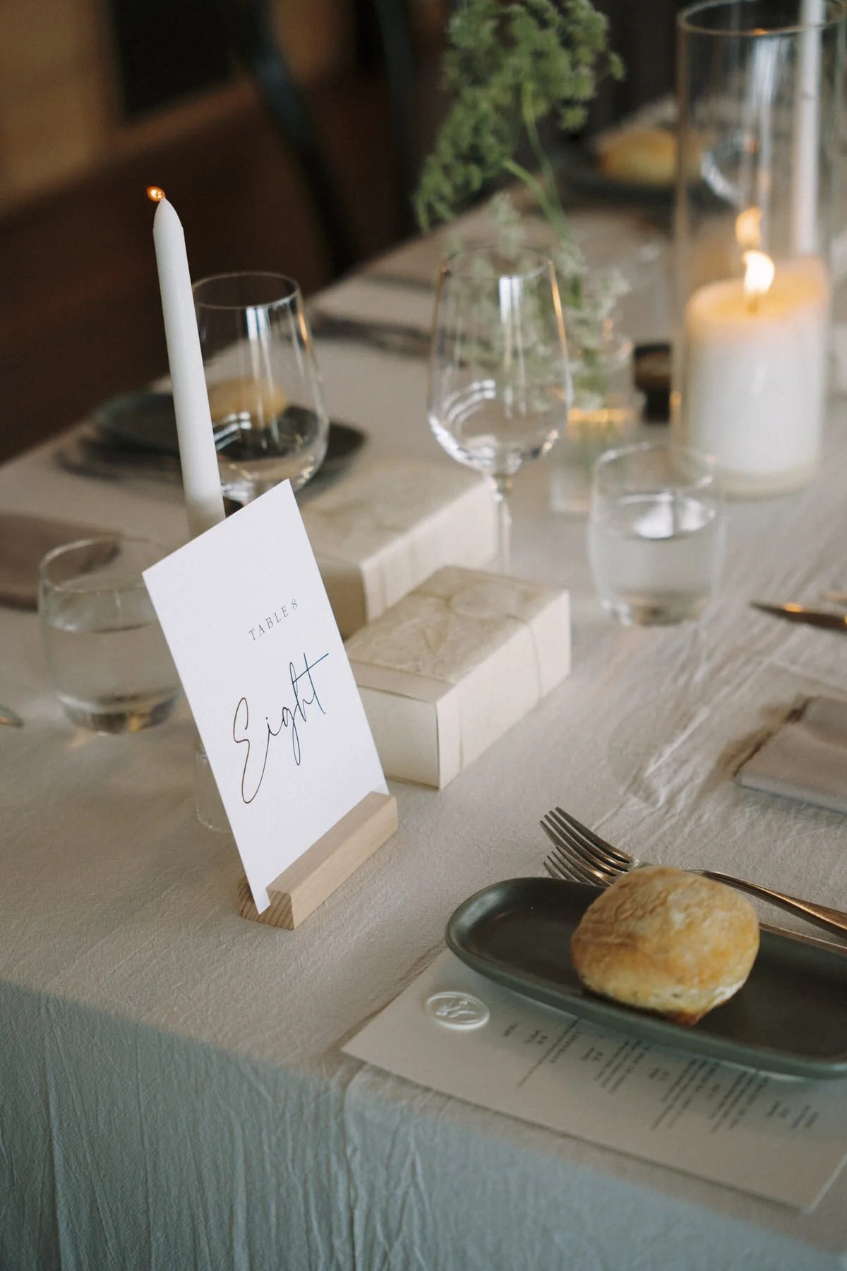 A table setting with a white tablecloth, a small sign that says 'Eat' in a wooden holder, a tall white candle, wine glasses, water glasses, candles, and a plate with a bread roll.