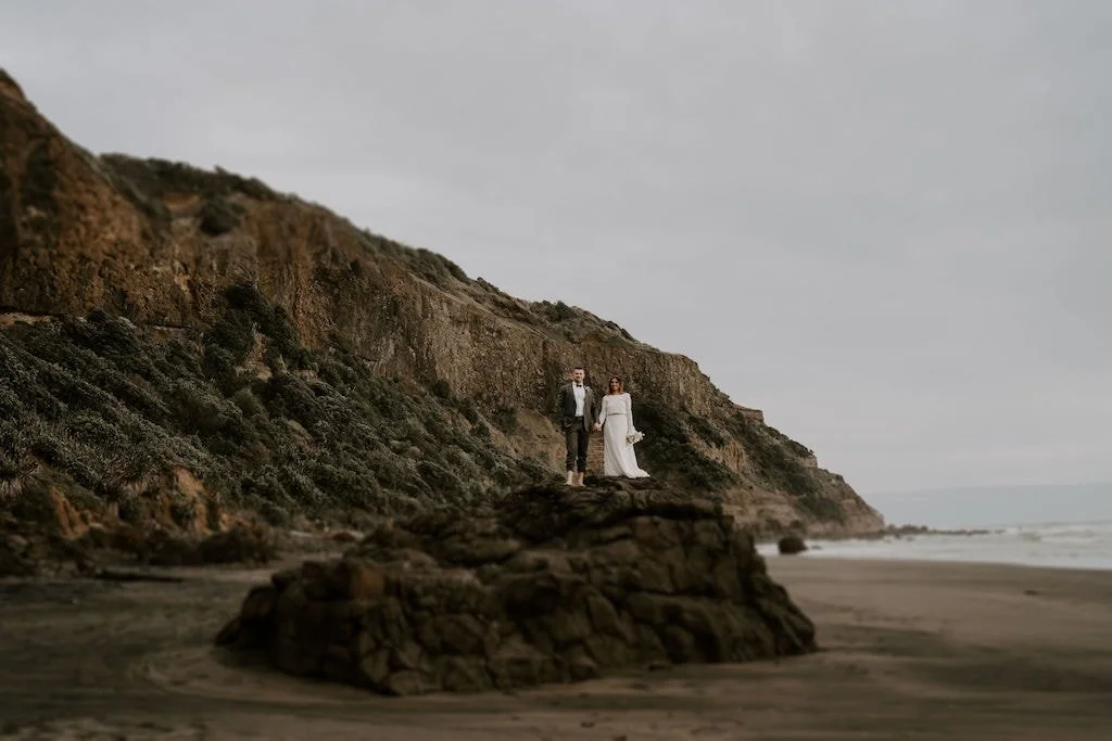 Newlywed couple embracing with a modern white and blush bridal bouquet on New Zealand's rugged West Coast.