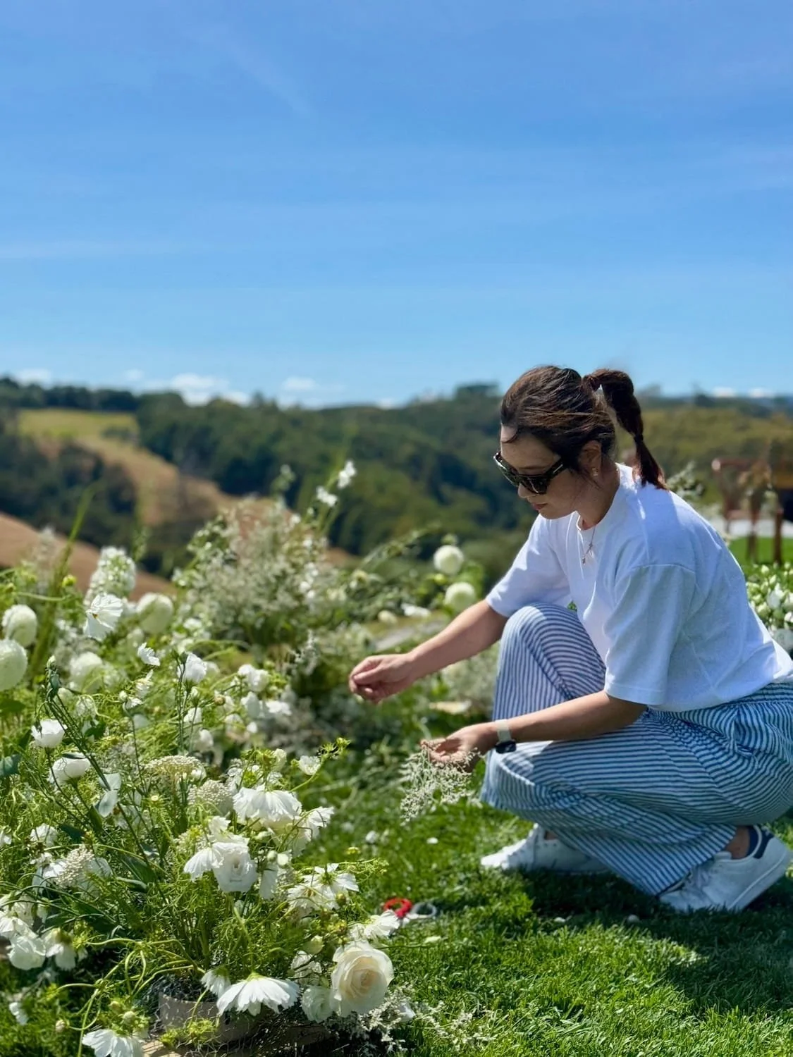 A woman wearing sunglasses, a white t-shirt, and striped pants crouches near white flowers on a sunny day with a blue sky and green rolling hills in the background.