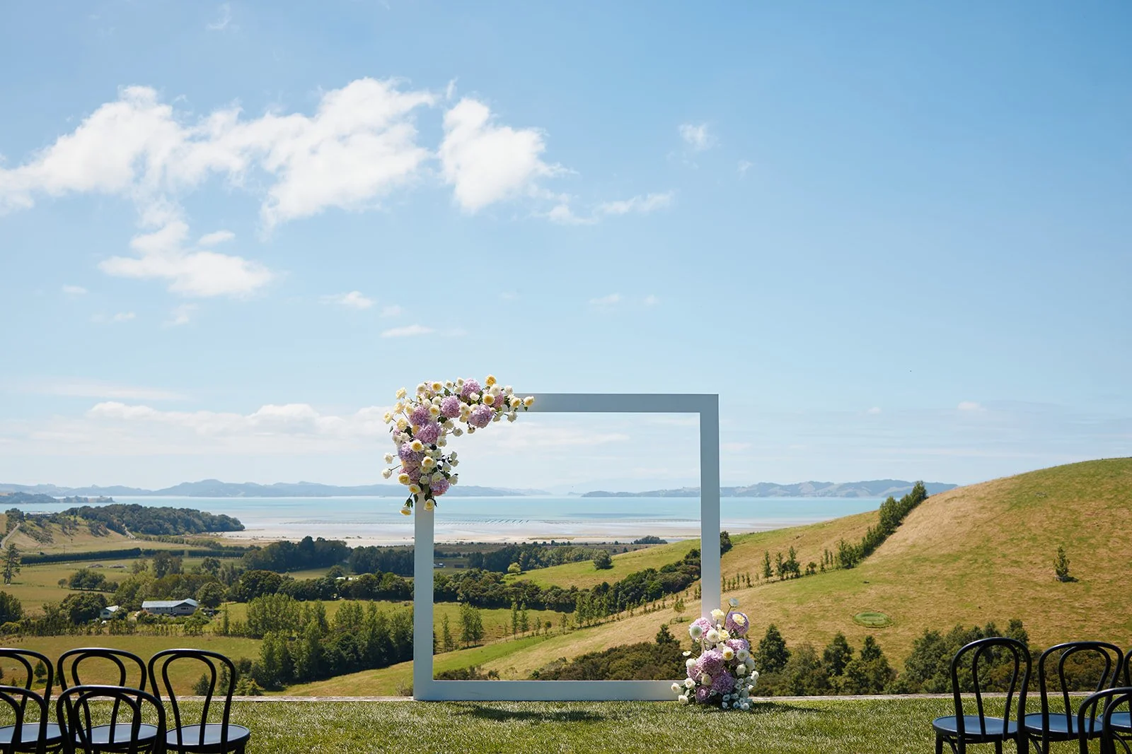 Summer wedding ceremony at Kauri Bay featuring a modern white floral frame overlooking the coast