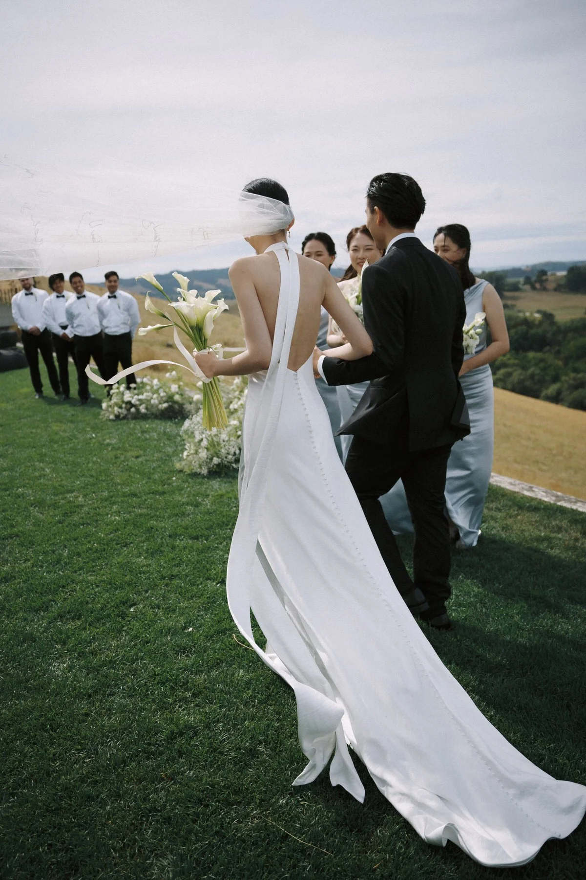 A bride wearing a white gown and veil holding a bouquet of white calla lilies, with a groom in a black suit, outdoors during a wedding ceremony with a backdrop of hills and a cloudy sky. Bridesmaids and groomsmen are standing in the background.