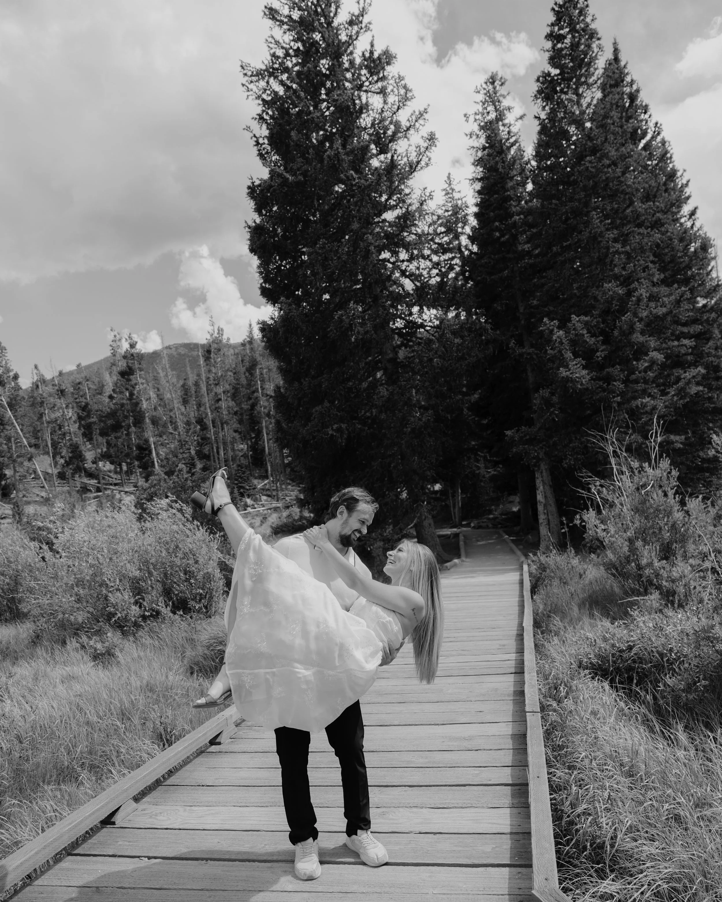 A man lifting a woman in a white dress on a wooden pathway in a natural outdoor setting with trees and bushes.