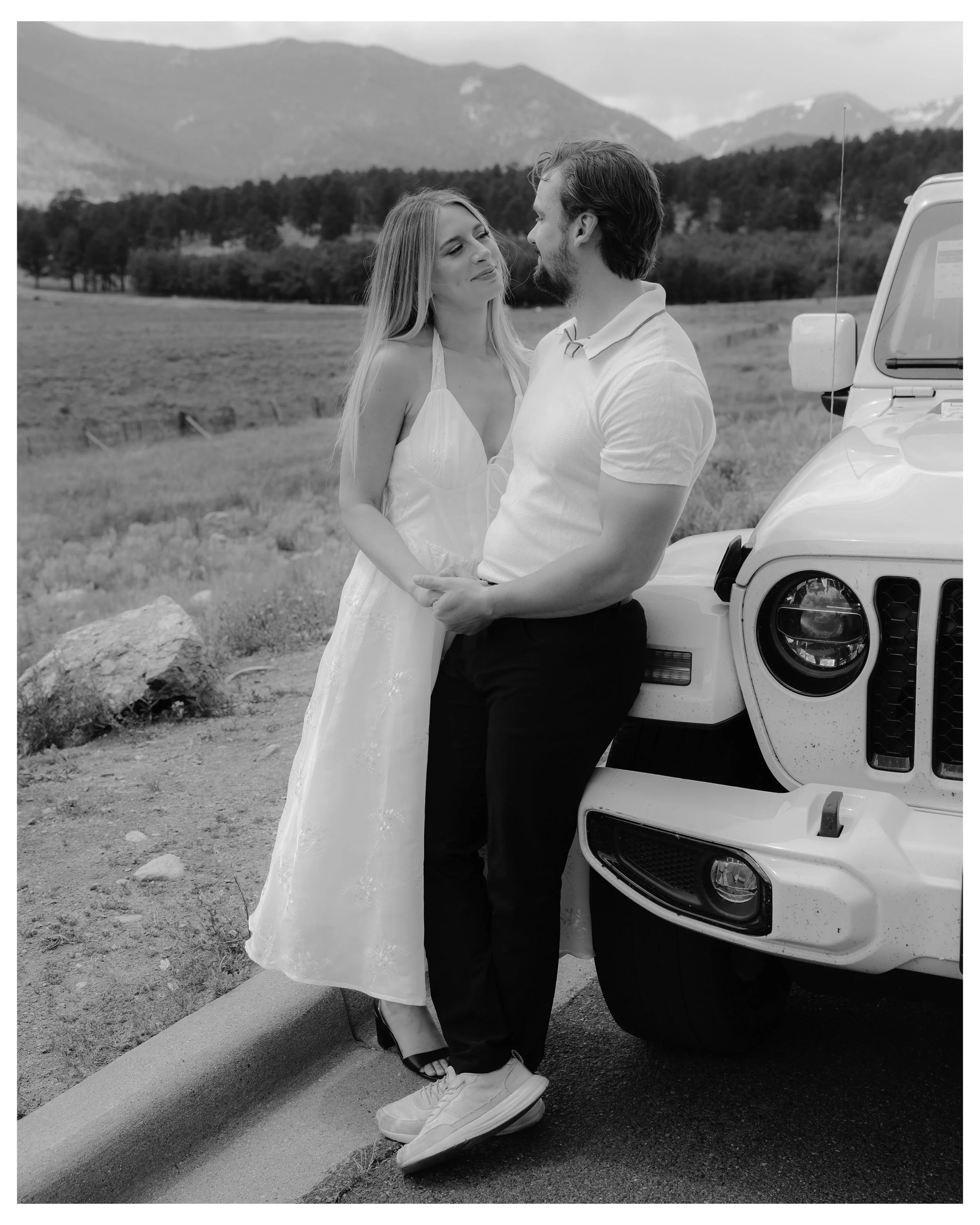 A couple standing by a white vehicle outdoors, with mountains and trees in the background.