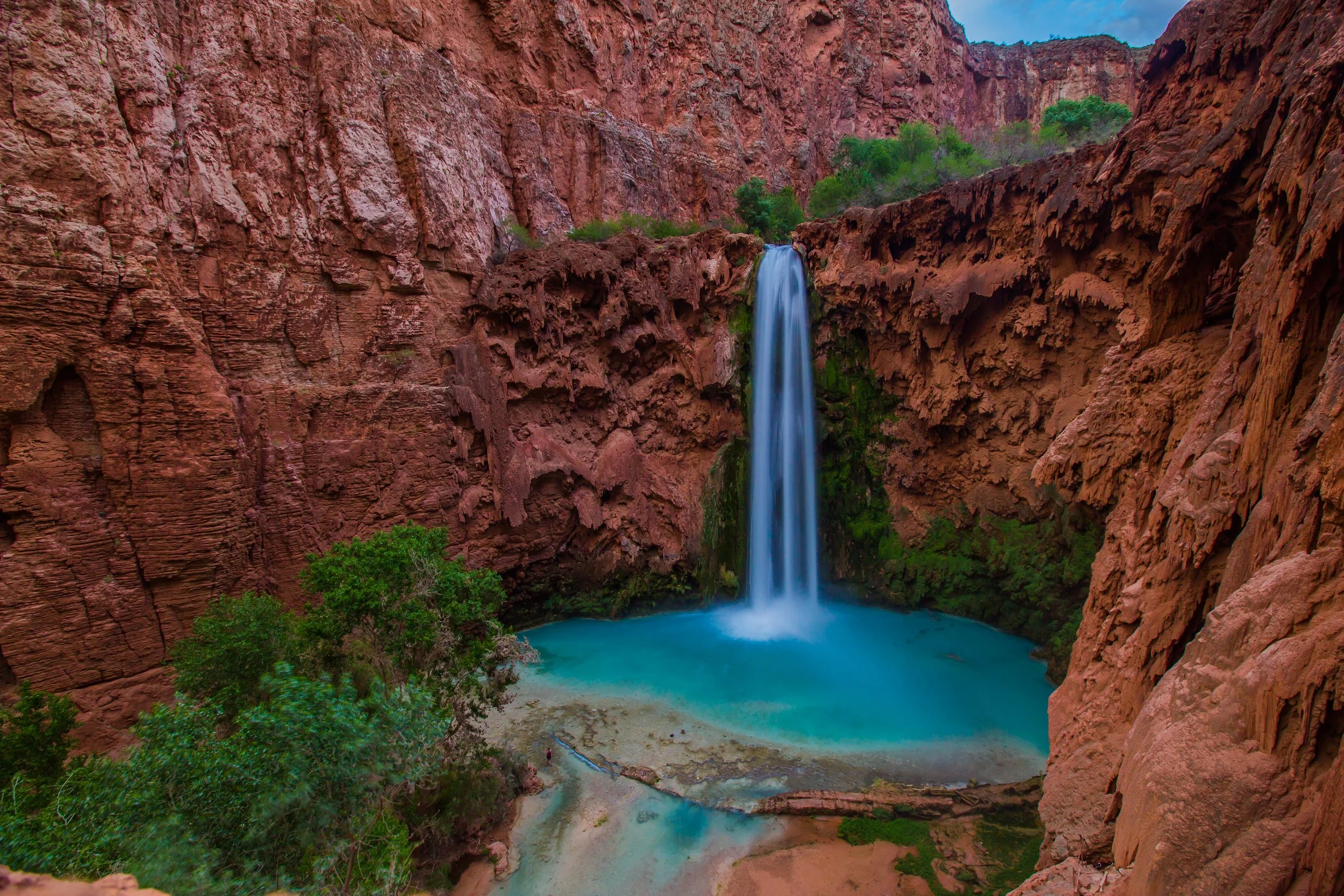 Turquoise water cascading over Mooney falls in Supai, Arizona, with red canyon walls reflecting sunlight