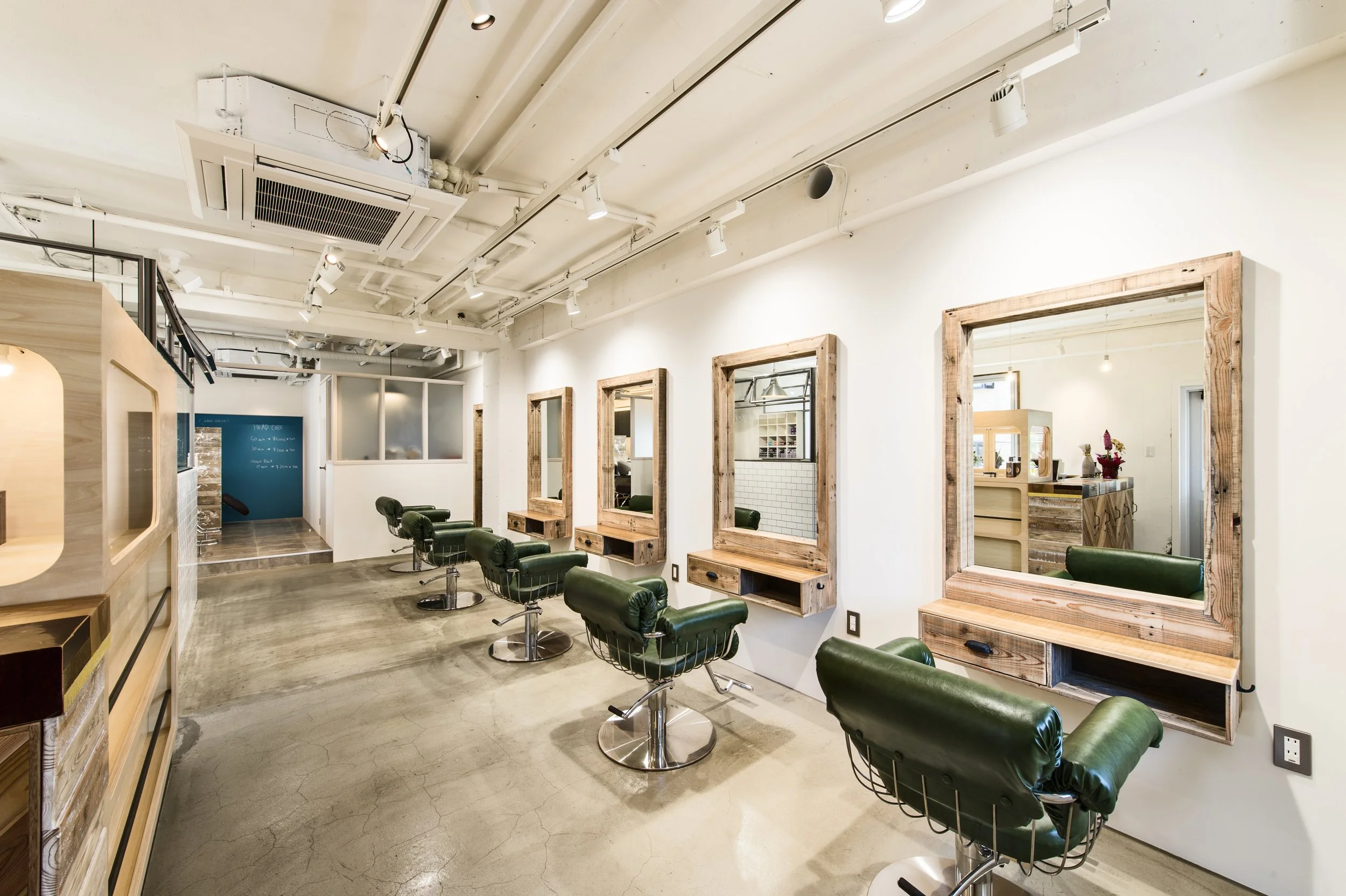 Interior of a modern hair salon with four styling stations, each with a large mirror framed in rustic wood, green leather chairs, and a small shelf. The salon has an industrial ceiling with exposed pipes and white walls, and polished concrete floors.
