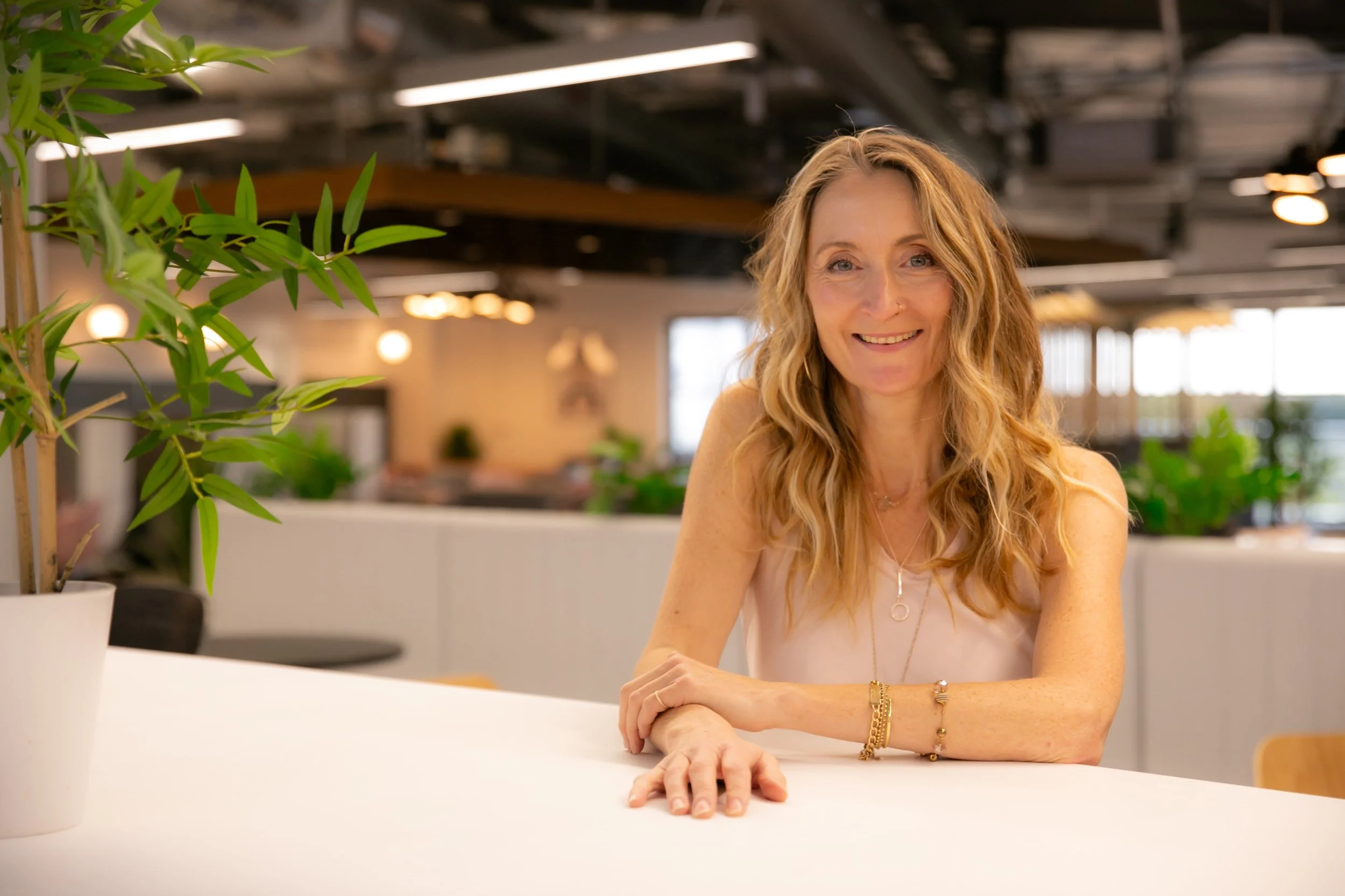 A woman with long, wavy blonde hair and a warm smile seated at a white table in a modern, brightly lit indoor space with plants and large windows.