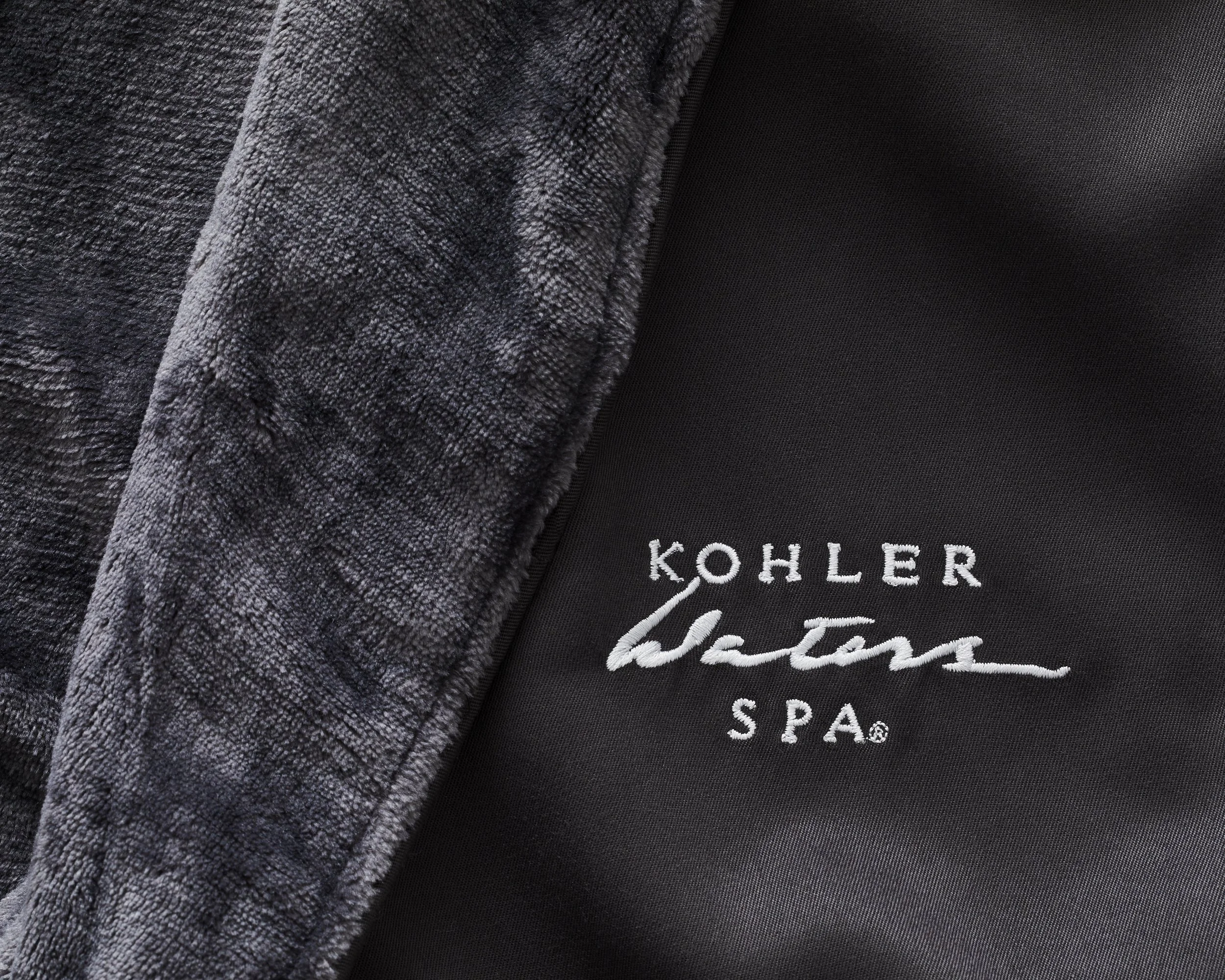 Close-up of a fabric with the embroidered logo 'Kohler Waters Spa' on black material, with part of a textured grey towel or cloth visible on the left.