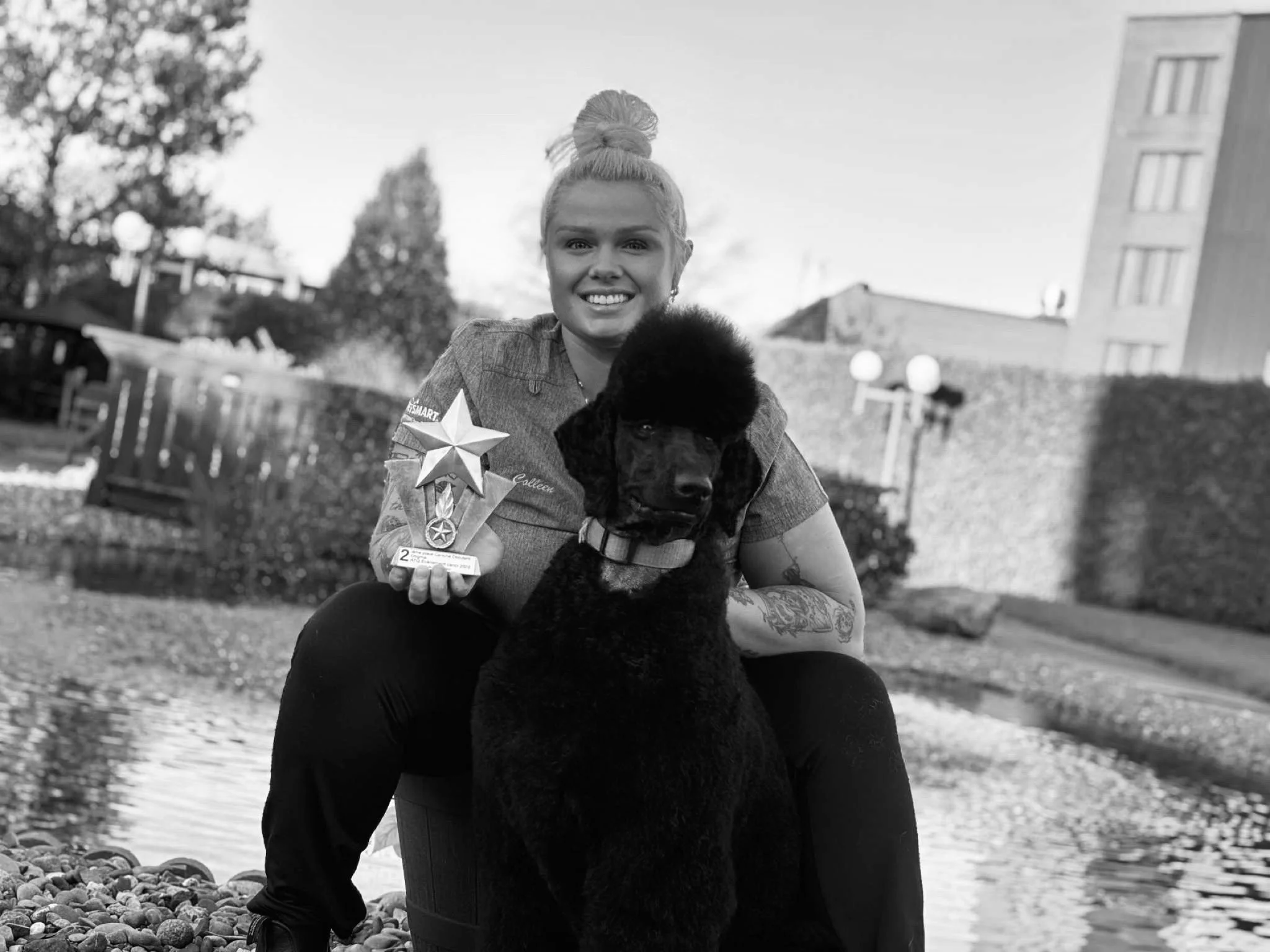 Black woman sitting on a barrel with a large curly-haired black dog, holding a shiny star-shaped award, outdoors near a body of water, with trees and buildings in the background.