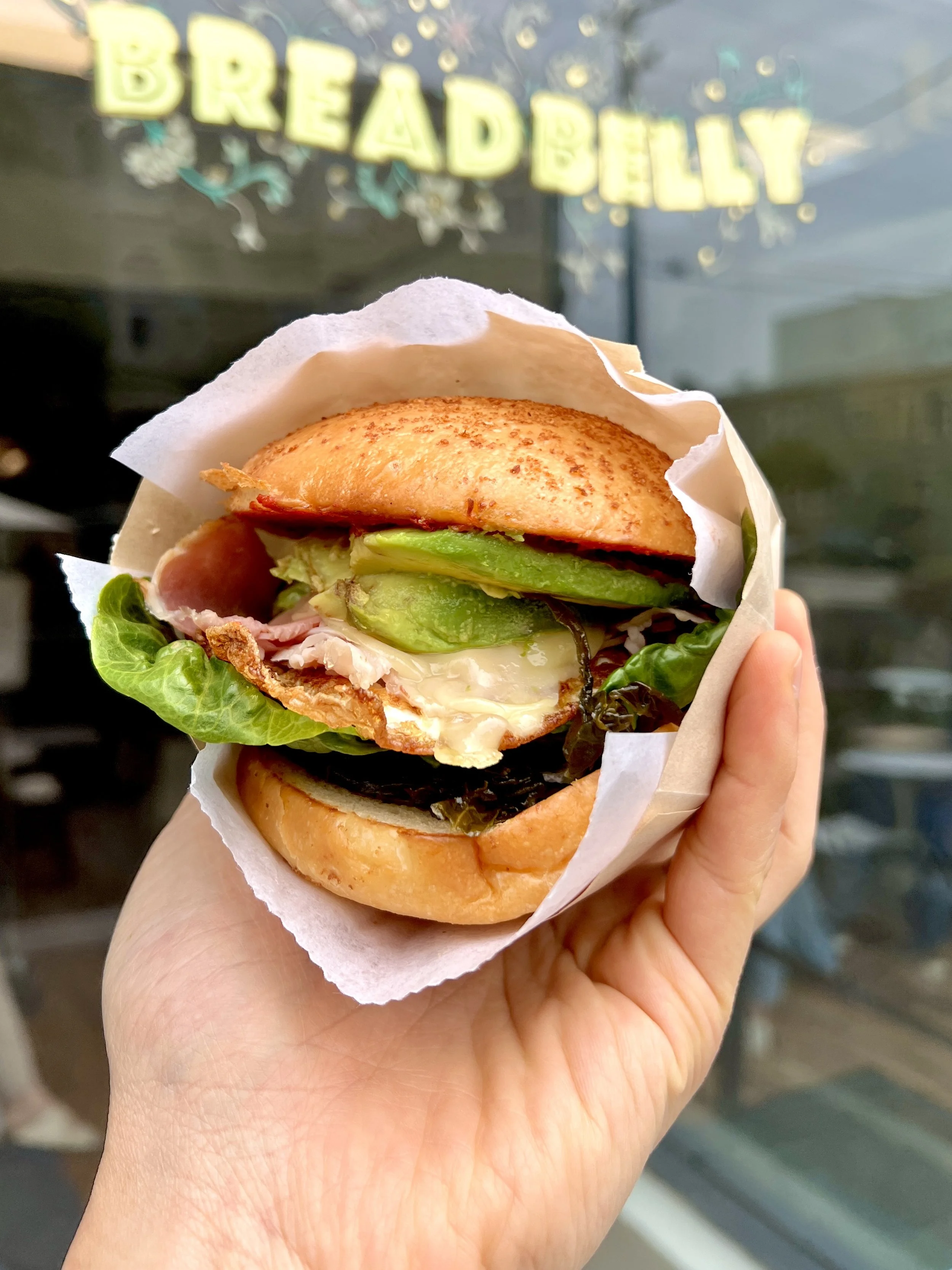 A hand holding a sandwich with lettuce, pickles, cheese, ham, and egg on a bun, wrapped in white parchment paper.
