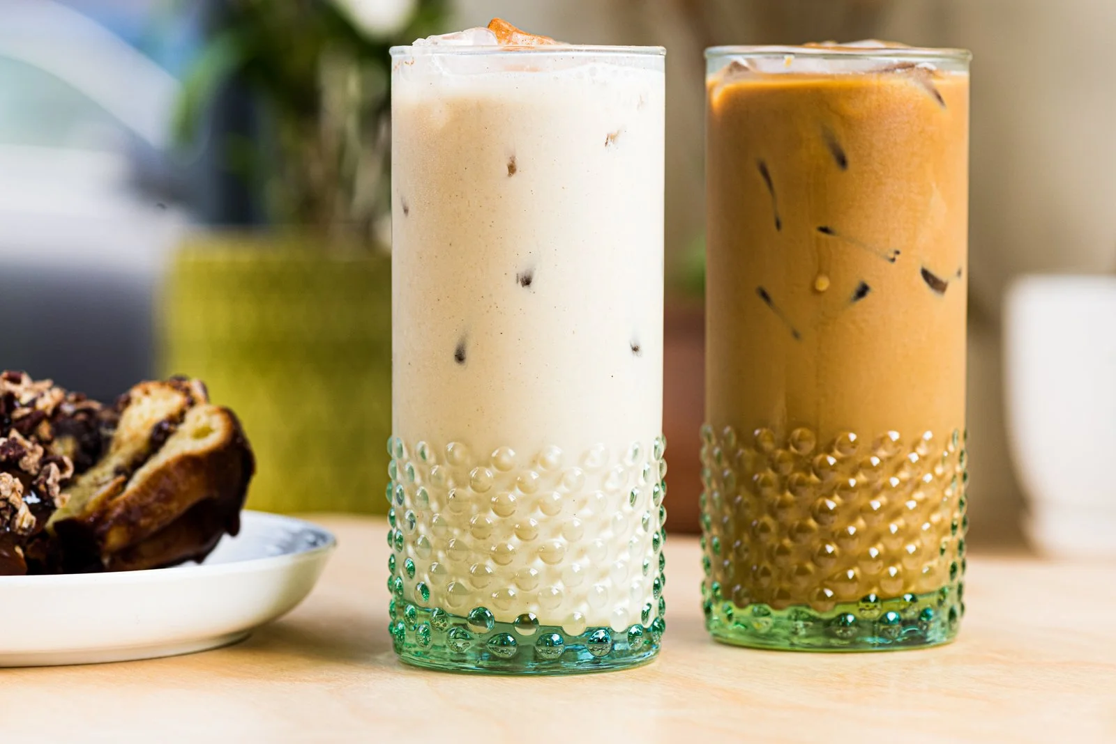 Two tall glasses of iced coffee beverages on a wooden surface. The left glass contains a light-colored iced coffee with ice cubes, and the right glass contains a darker iced coffee with ice cubes. There is a partially visible plate with a dessert on 