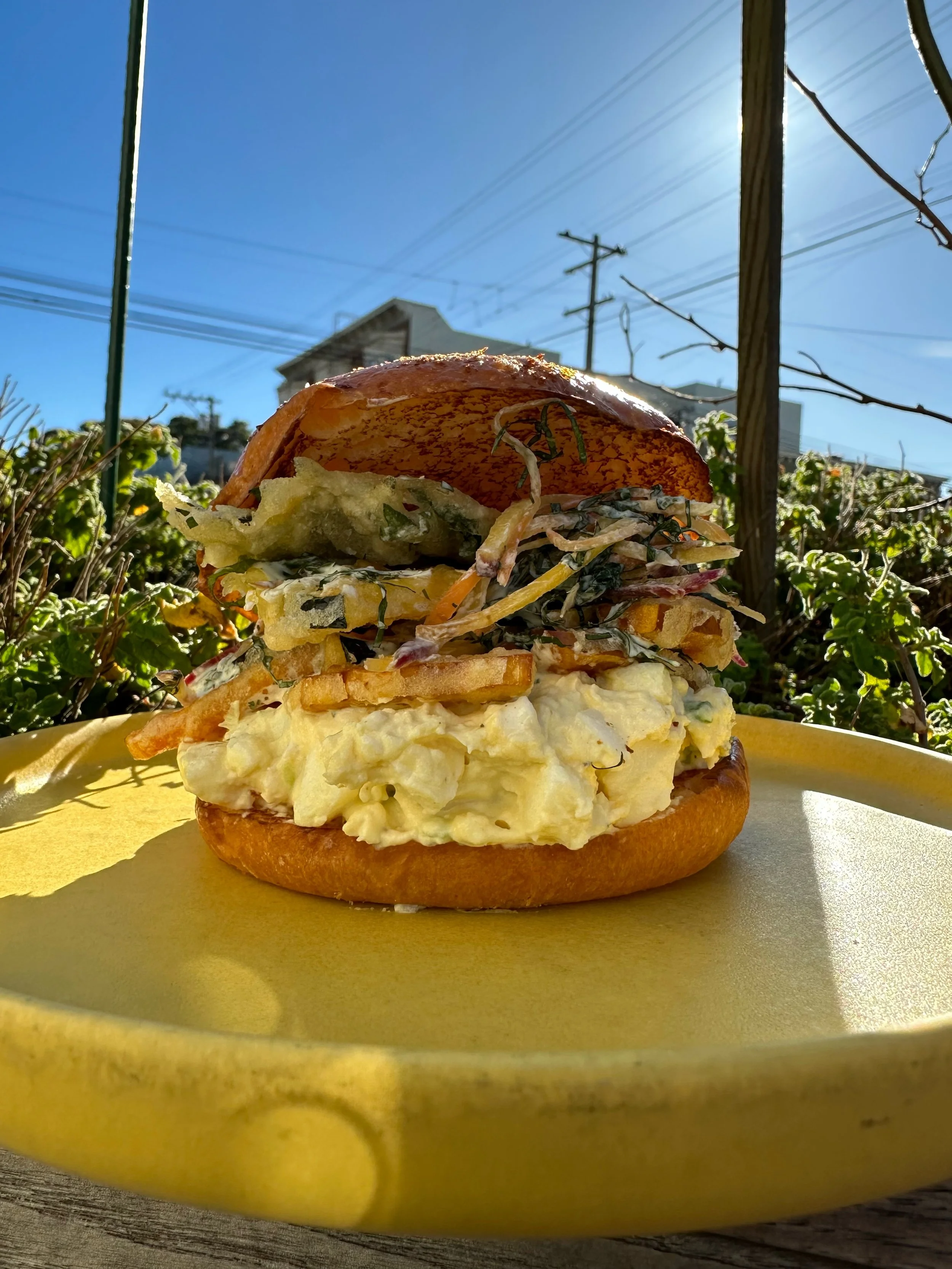 Close-up of a egg salad sandwich in a bun outdoors on a yellow plate with a garden.