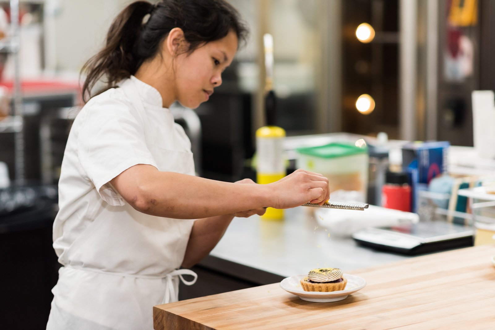Chef Katherine Campecino in a white chef's coat is decorating a tart with icing or frosting in a commercial kitchen.
