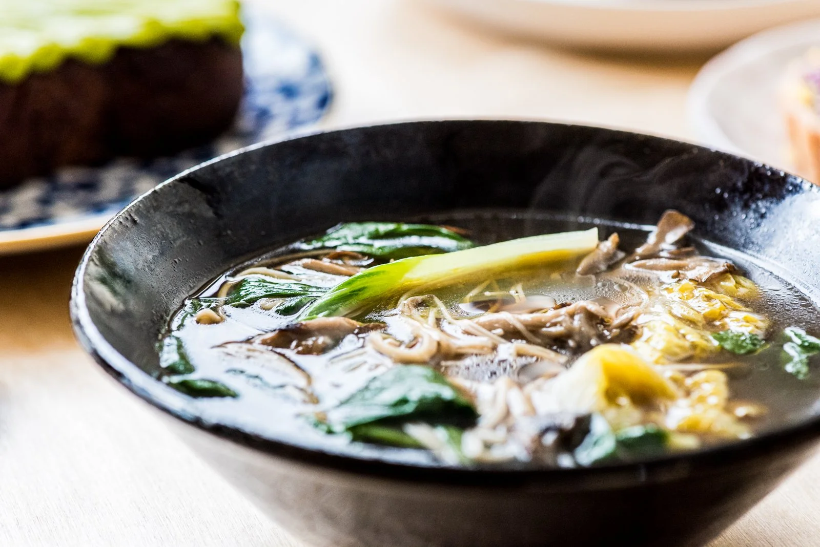 A black bowl of hot soup with greens and noodles on a wooden table, with blurred dishes in the background.