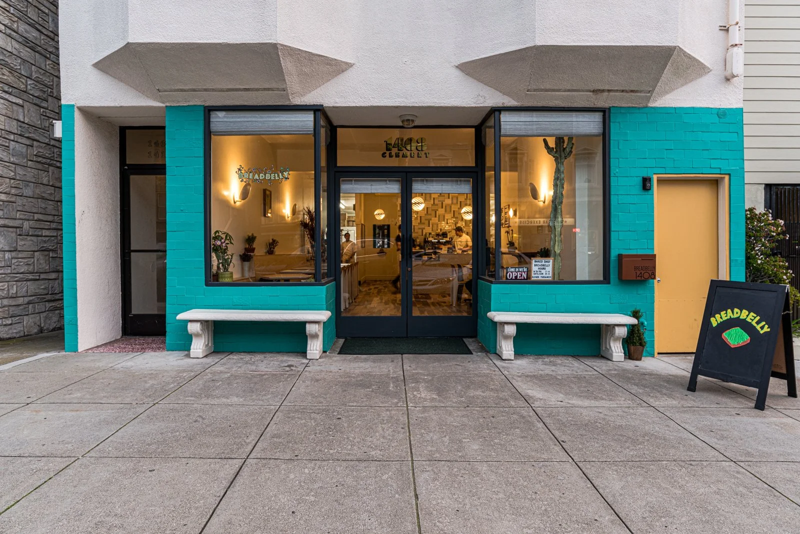 Front view of a small restaurant called Breadbelly with turquoise and yellow exterior, large glass windows, two white benches outside, and a black chalkboard sign indicating it is open.